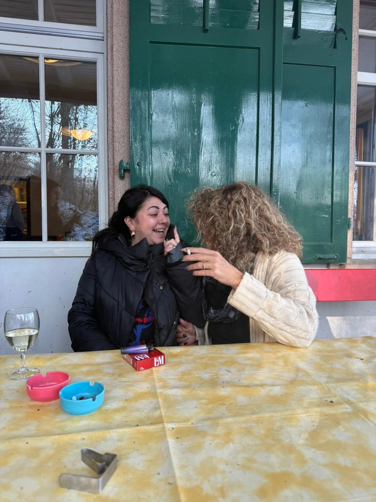 Zwei Frauen lachen zusammen an einem Tisch im Freien. Eine Frau trägt eine schwarze Jacke, die andere eine beige Strickjacke. Es gibt einen Wein- oder Sektglas, einen Zigarettenpack, zwei bunte Aschenbecher und einen Messer auf dem Tisch.