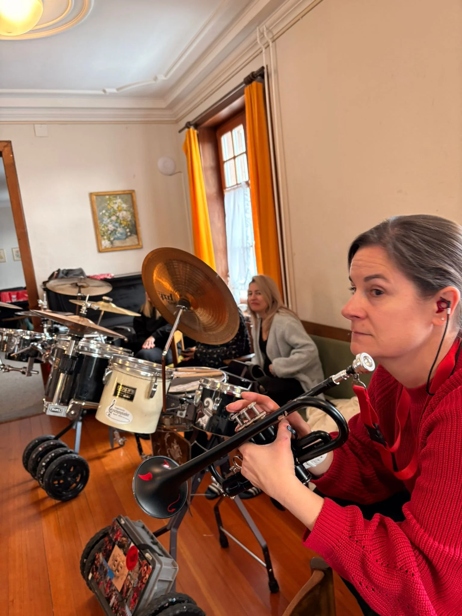 Musikerinnen während eines Proben- oder Auftritts in einem Raum mit Holzfußboden und Fenstern mit gelben Vorhängen, die eine Trompete und ein Cajón spielen.