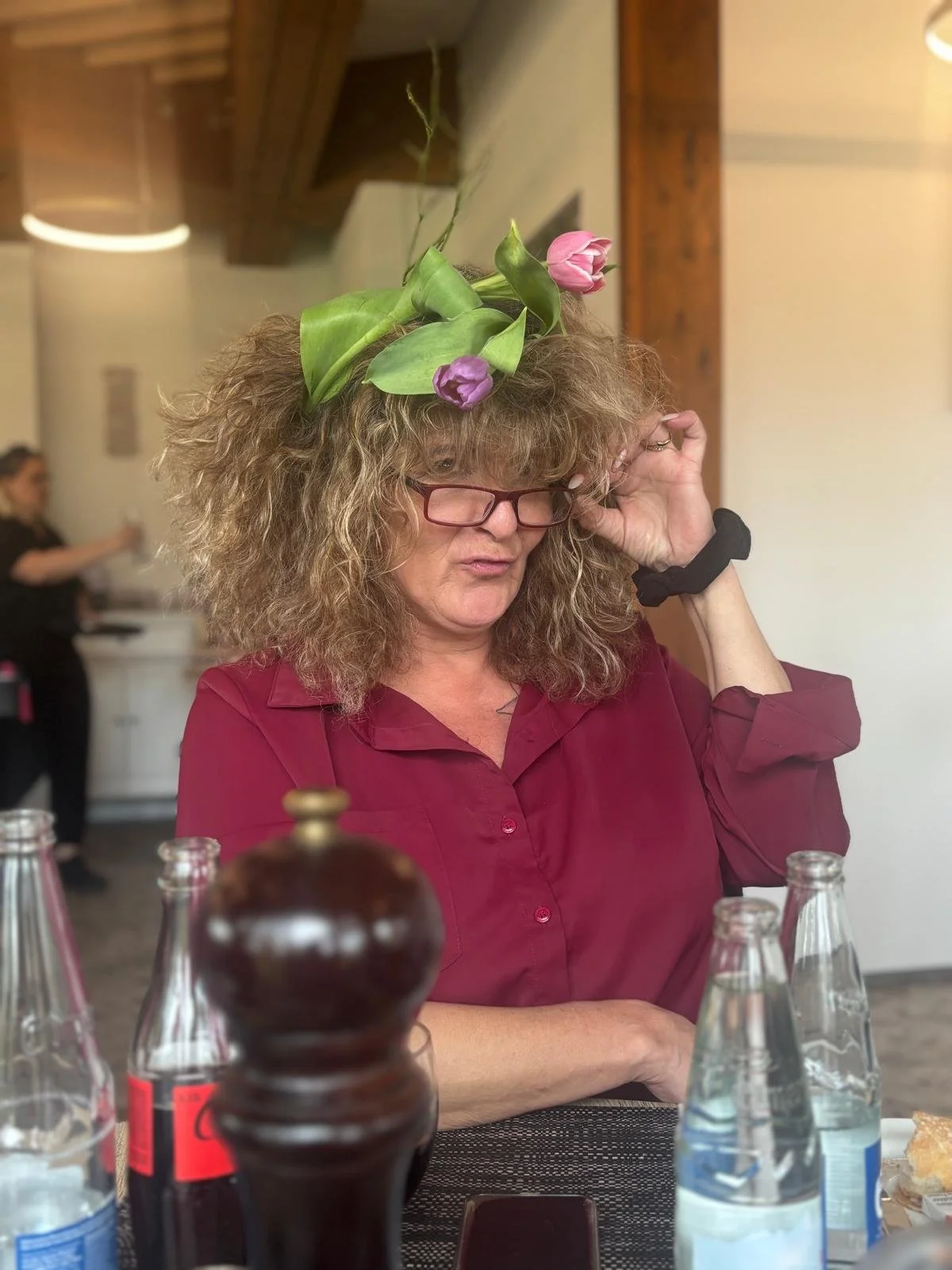 Frau mit großen, lockigen Haaren, trägt eine Fliederblume auf dem Kopf und liest ein Buch, sitzt an einem Tisch.