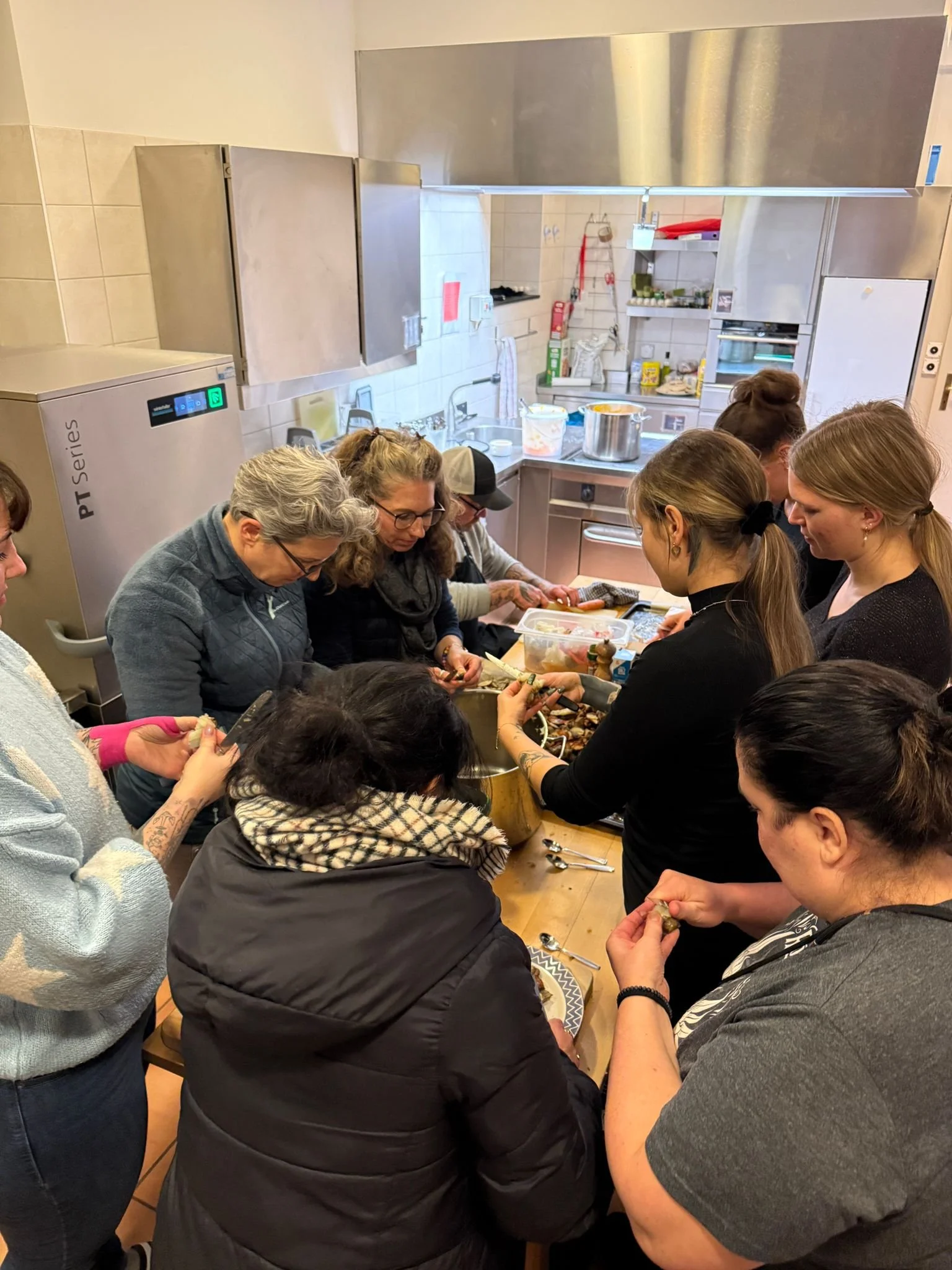 Mehrere Frauen bereiten gemeinsam in einer Küche eine Mahlzeit zu. Sie sitzen und stehen um einen Holz Tisch, während sie Lebensmittel schneiden und zubereiten.