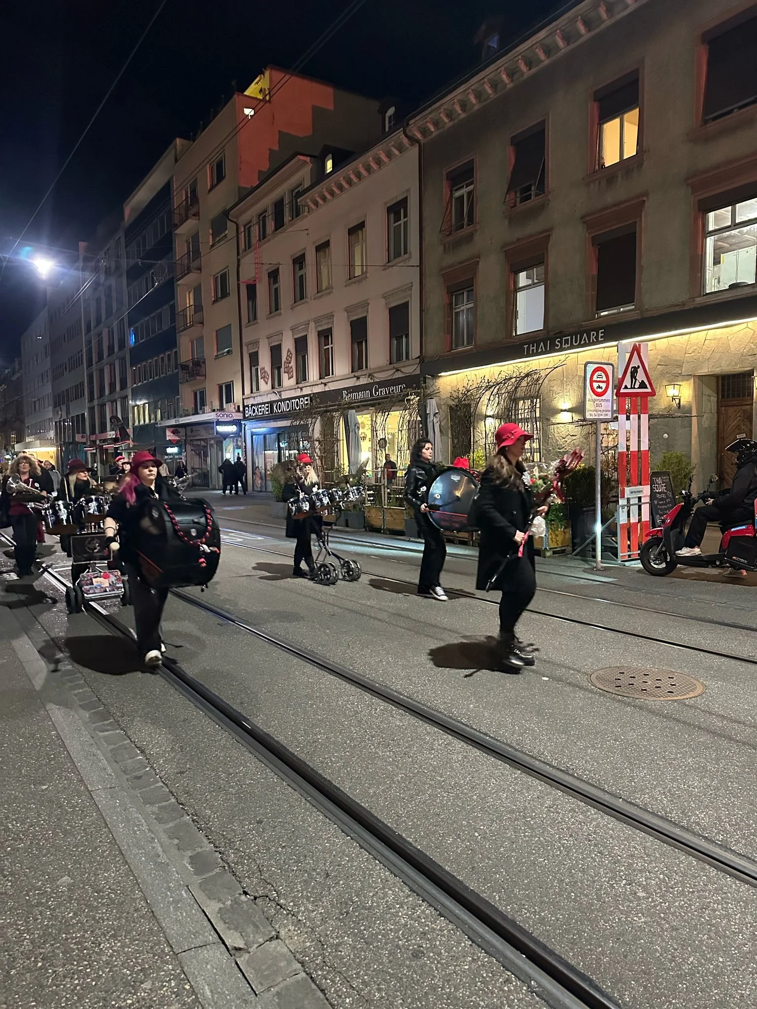 Menschen in einer Stadtstraße bei Nacht, einige mit roten Hüten und Regenschirmen, die auf Tramgleise gehen, mit Geschäften im Hintergrund.