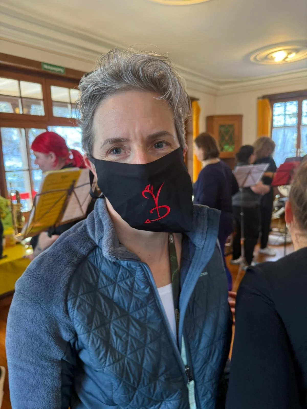 Eine Frau mit kurzgrauem Haar trägt eine schwarze Maske mit rotem Symbol. Im Hintergrund spielen Musiker mit Notenständern im Raum.