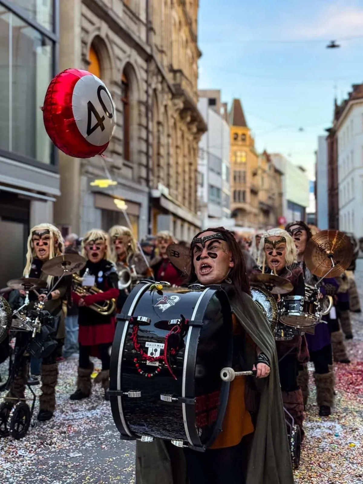 Leute in Kostümen bei einer Parade auf der Straße, mit Musikinstrumenten und Luftballons, im Hintergrund historische Gebäude, bunte Konfetti auf dem Boden.
