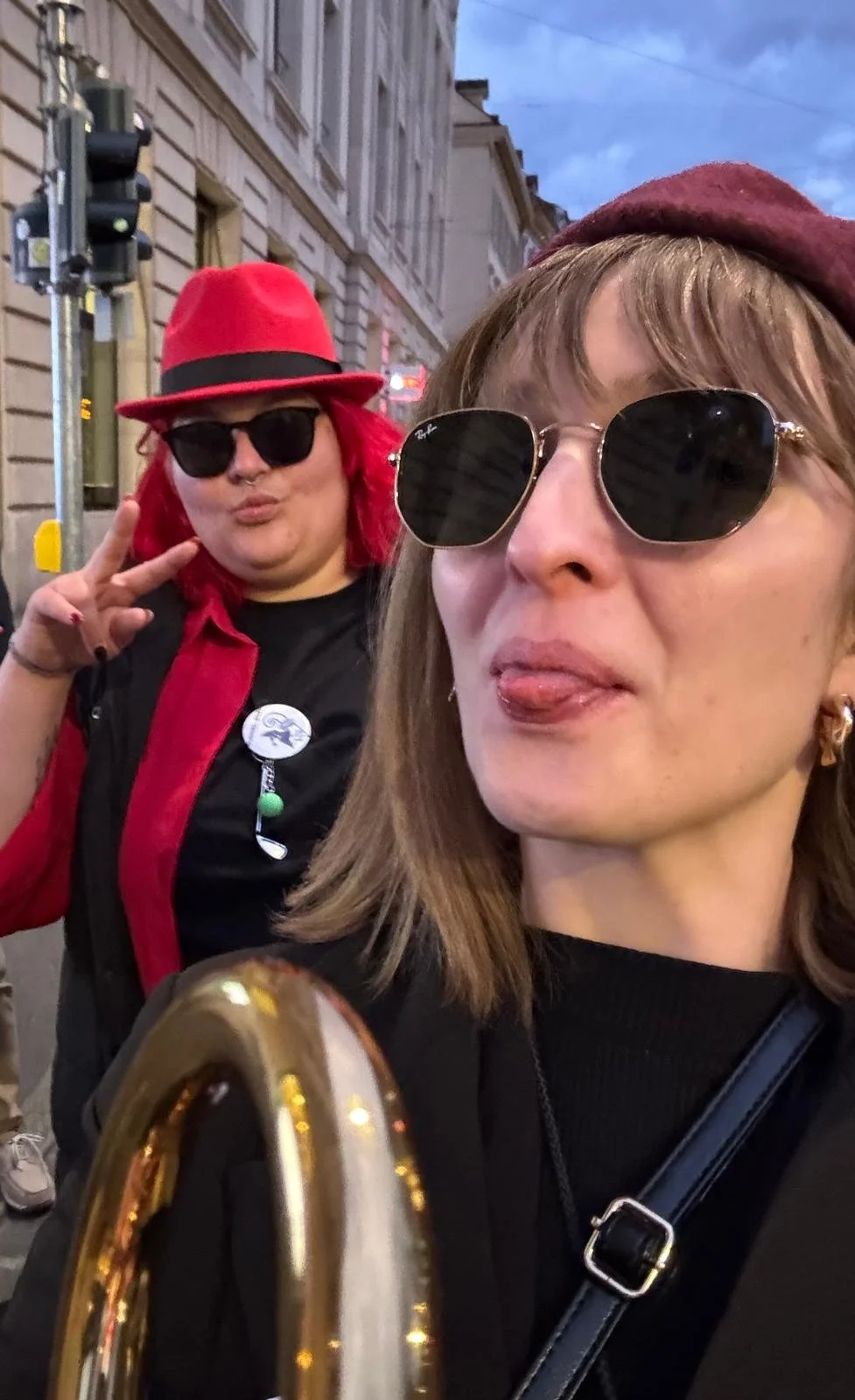 Zwei Frauen tragen Sonnenbrillen und machen Selfies in einer urbanen Straße, eine mit rotem Hut und die andere mit rot gefärbtem Haar und schwarzem Shirt.