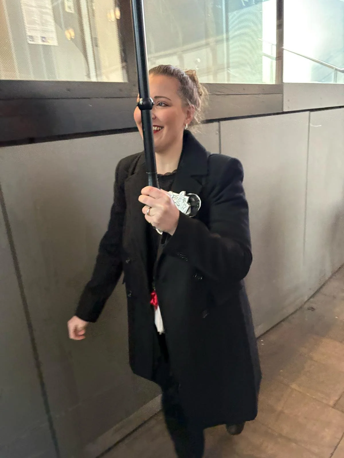 Eine lachende Frau mit einem schwarzen Mantel hält einen schwarzen Regenschirm, während sie durch eine städtische Gegend läuft.