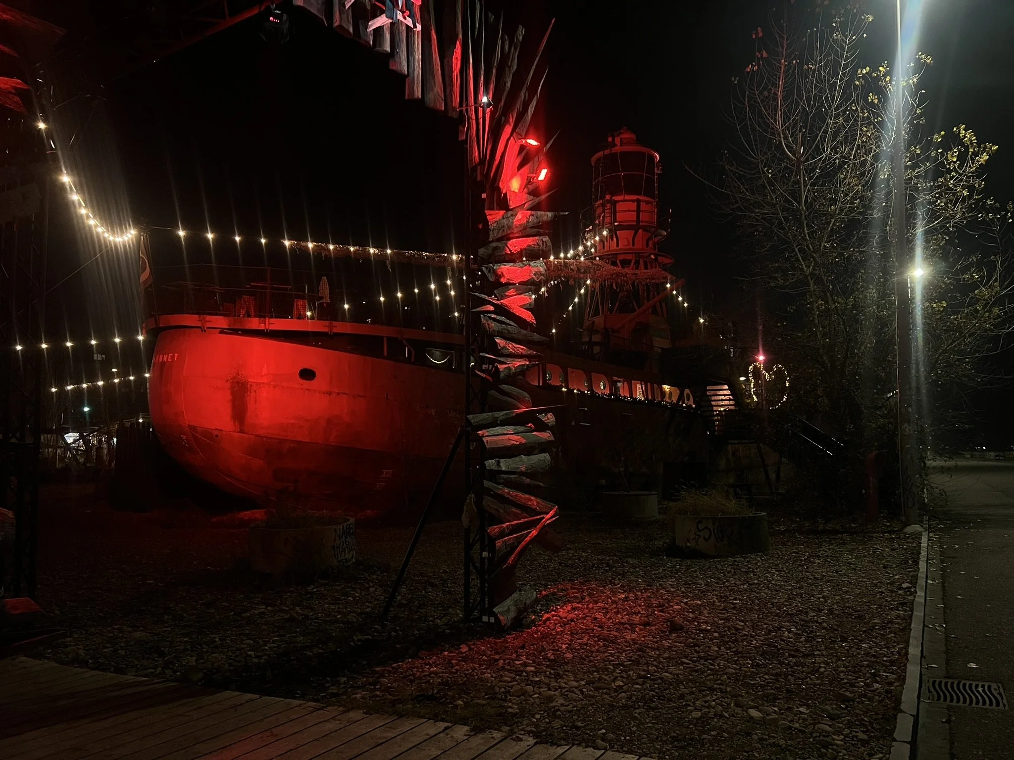 Ein altes, rot gestrichenes Schiff auf Land, nachts beleuchtet mit Lichterketten, umgeben von Bäumen und Laternen. Es ist dunkle Atmosphäre mit künstlichem Licht und Schatten.