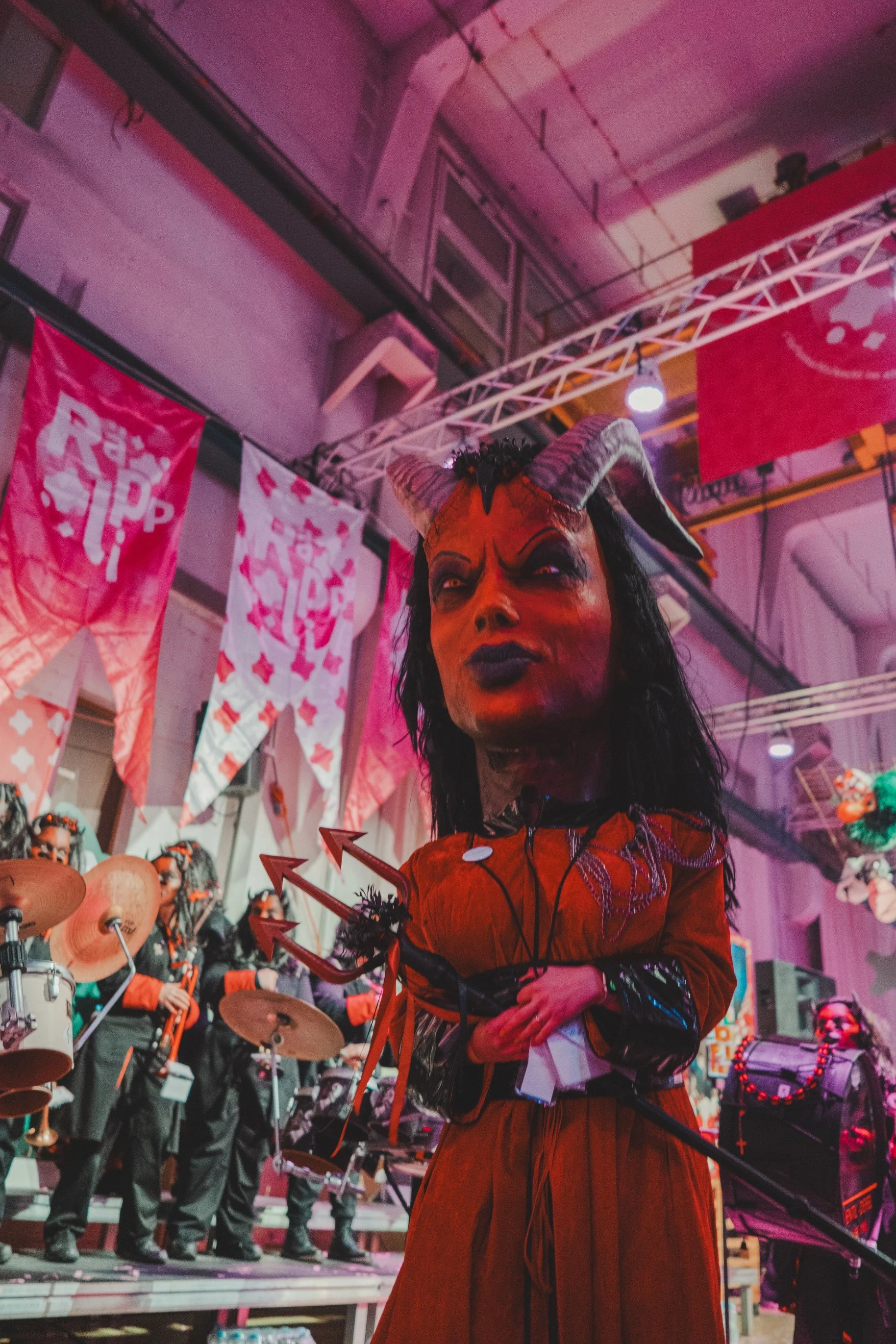 Eine große Figur mit dämonischem Gesicht, Hörnern und schwarzen Haaren, in einer roten Kleidung, in einem festlich geschmückten Innenraum mit einer Band im Hintergrund.