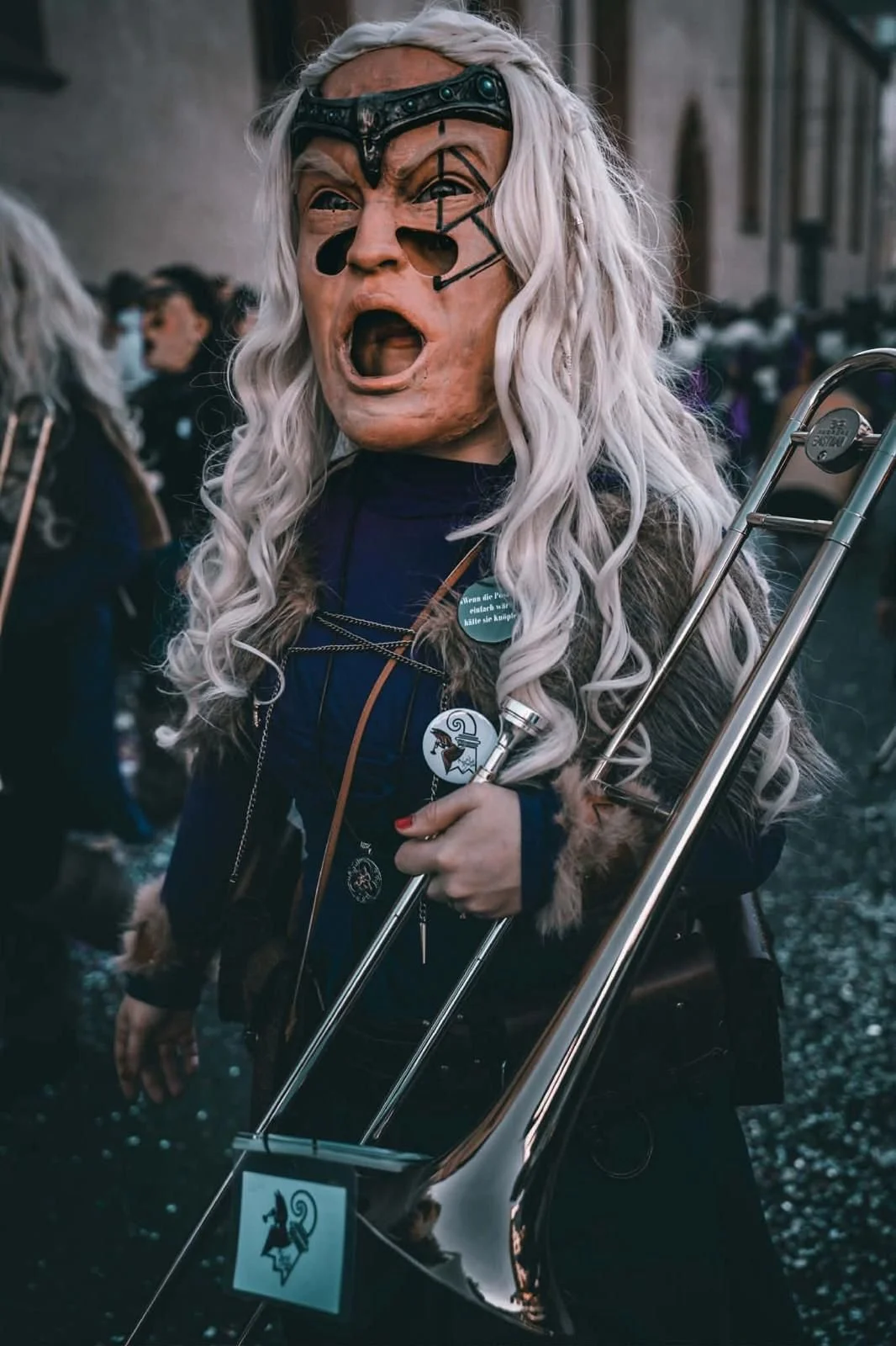 Person trägt eine große Maske mit weißen Locken und schwarzen Elementen, hält eine Bassgitarre, trägt Jacke mit Pins, in einer Menschenmenge bei einer Veranstaltung.