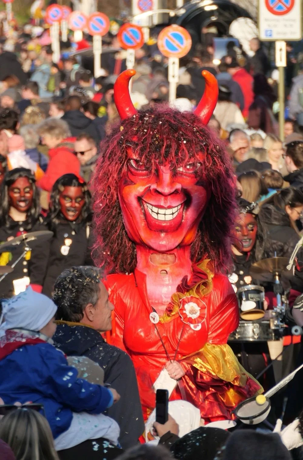 Eine große Person in einem roten Teufelskostüm mit roten Hörnern, einem maskierten, lachenden Gesicht und langem, lockigem Haar, umgeben von jemandem mit einem Smartphone und einer Gruppe von Menschen, die an einem Straßenfest oder Karneval teilnehme