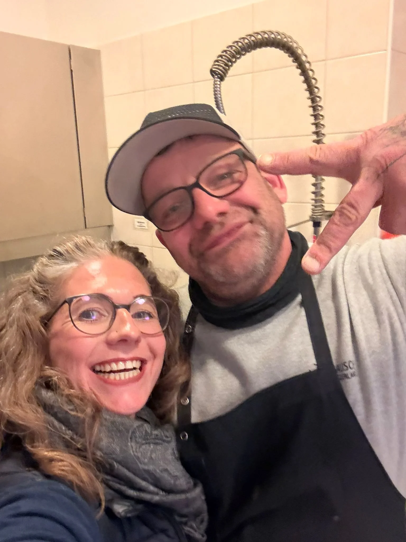 Zwei Menschen, eine Frau und ein Mann, heben die Hand und machen ein Peace-Zeichen, während sie in einer Küche stehen. Der Mann trägt eine Kochschürze und eine Baseballkappe, beide sehen glücklich aus.