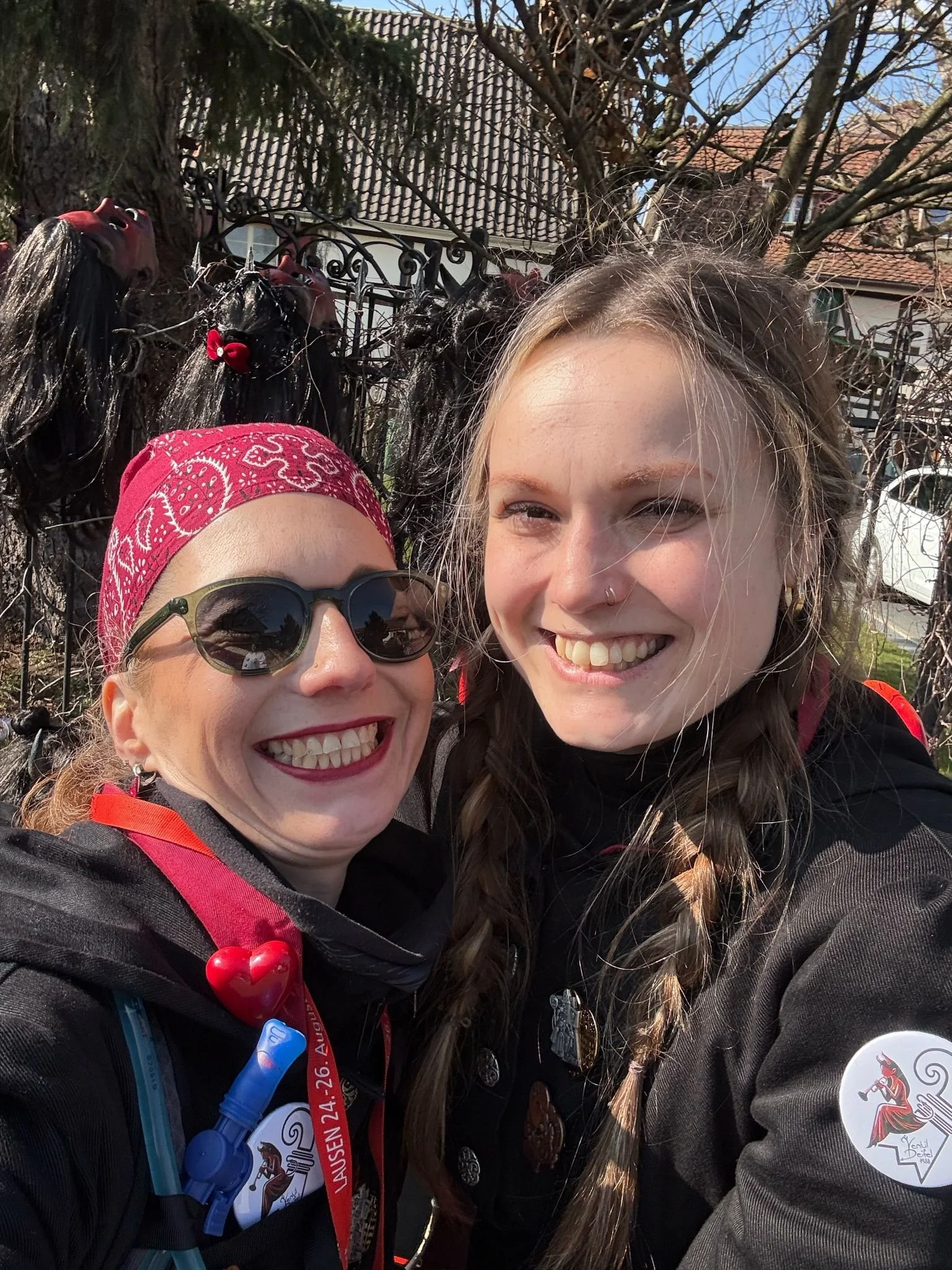 Zwei lachende Frauen, eine trägt eine pinke Bandana und Sonnenbrille, die andere hat einen Nasenring und trägt eine schwarze Jacke. Im Hintergrund ein Zaun mit schwarzen Hühner-Attrappen, eine Häuserwand und Bäume bei Sonnenschein.