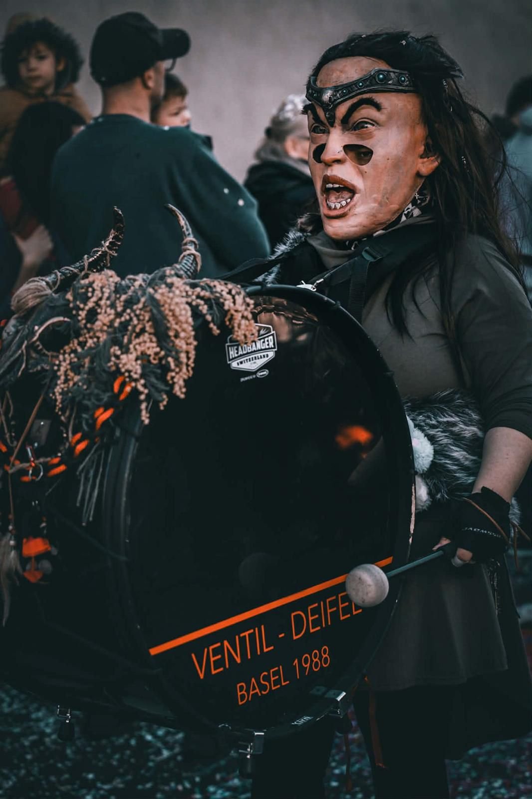 Eine Person in einem Kostüm mit einem maskenhaften Gesicht und Hörnern, die eine Trommel trägt, bei einer Veranstaltung. Im Hintergrund sind einige Menschen zu sehen.