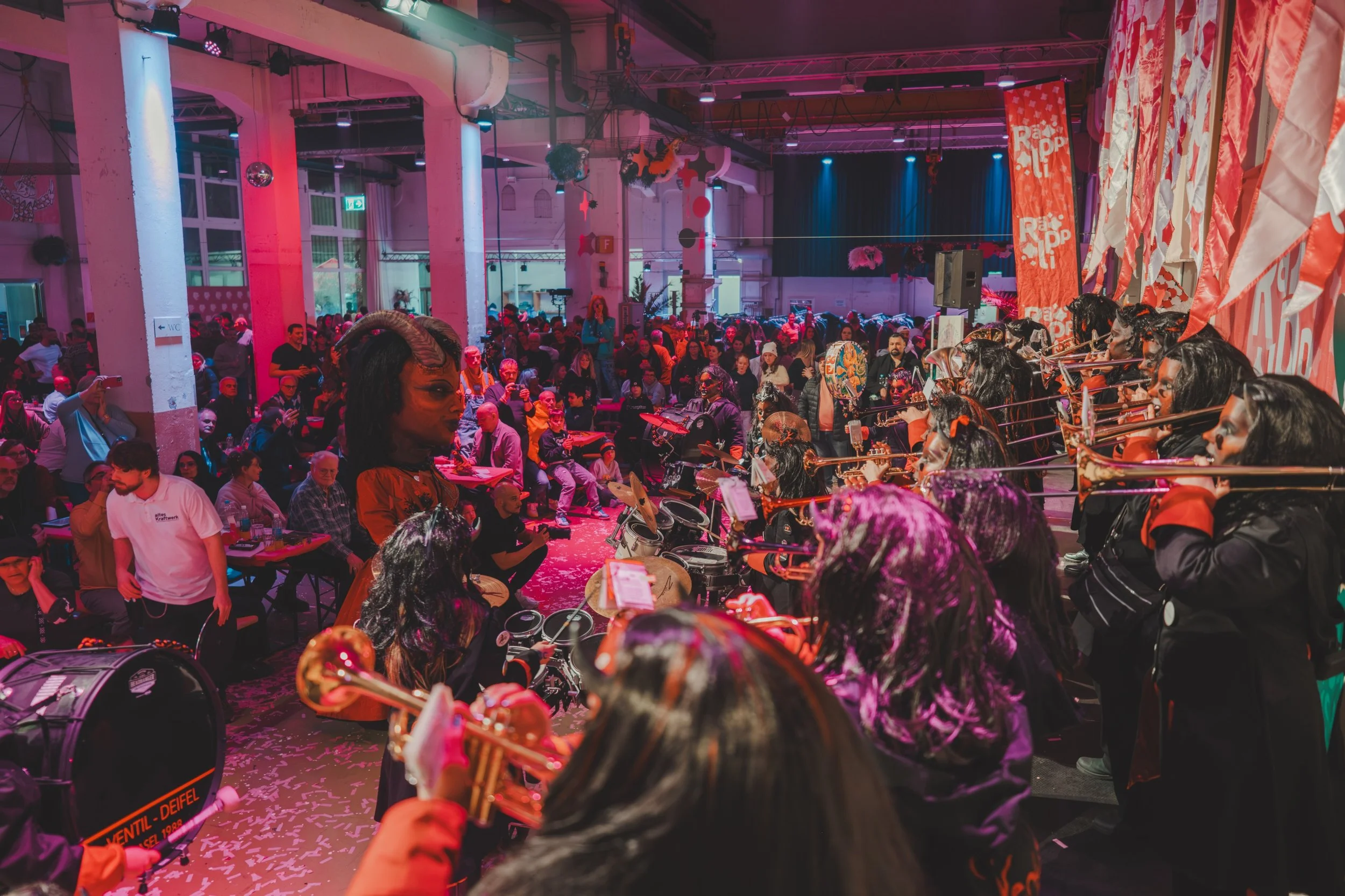 Menschen bei einem Indoor-Musik-Event, darunter ein Orchester mit Trompeten, im Hintergrund Publikum und bunte Beleuchtung, festliche Dekorationen