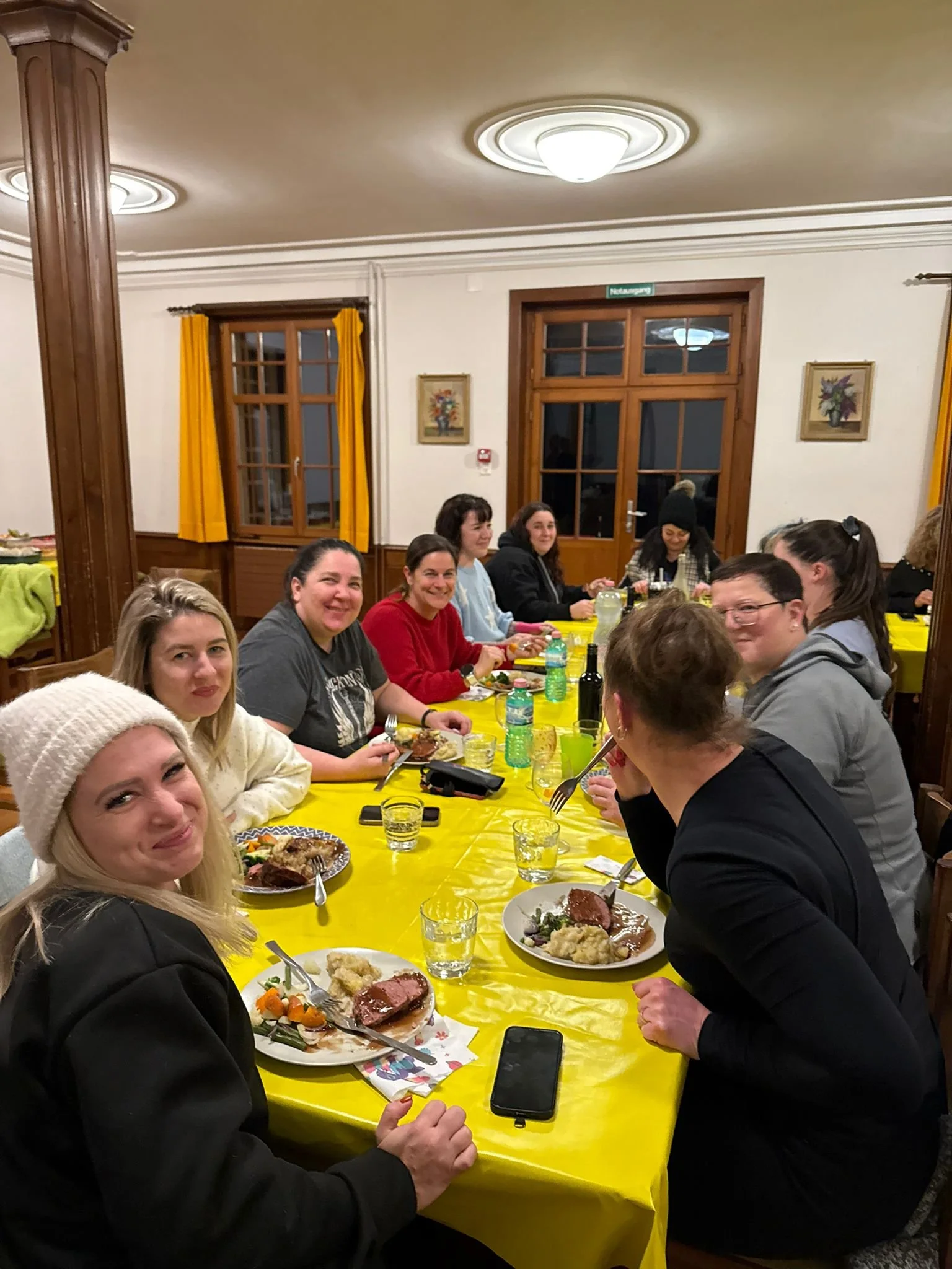 Menschen sitzen bei einem gemeinsamen Essen an einem langen Tisch mit gelber Tischdecke in einem festlich dekorierten Raum.
