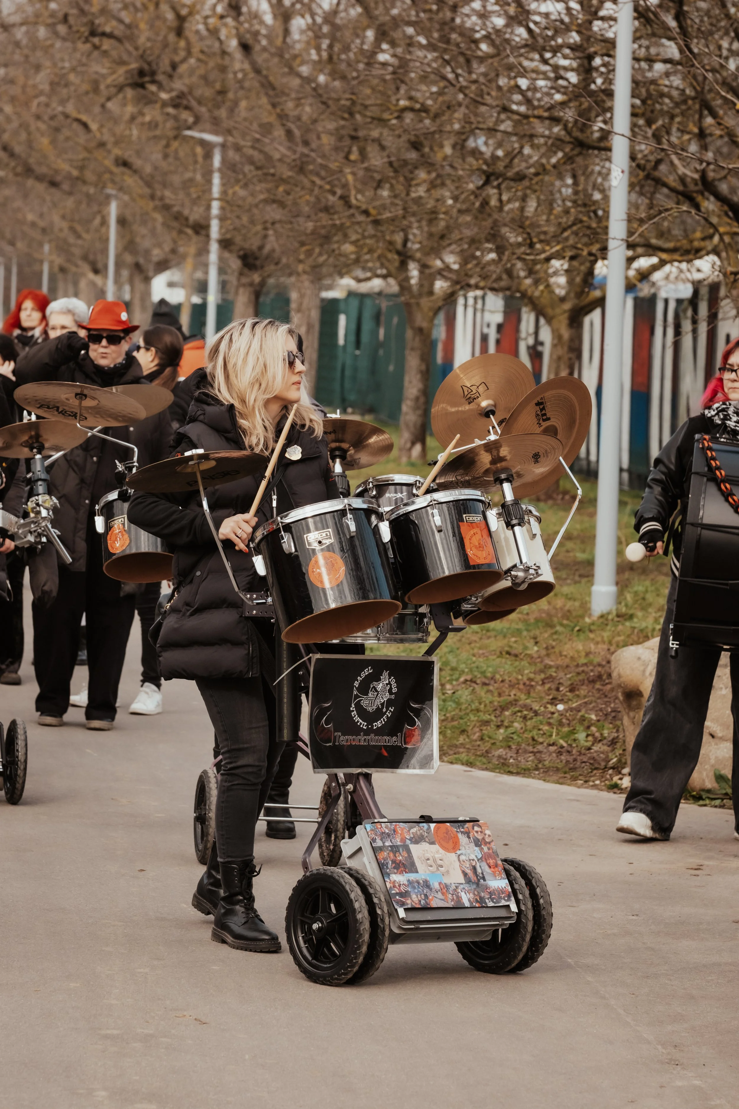 Eine Frau spielt auf einer tragbaren Trommel im Freien während eines Umzugs, umgeben von mehreren Menschen, einige tragen rote Hüte und dunkle Kleidung, im Hintergrund sind Bäume ohne Blätter.