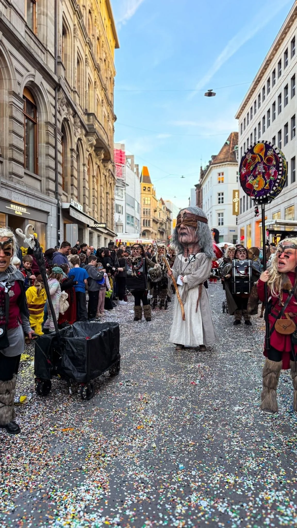 Faschingumzug auf einer Stadtstraße mit Menschen in Kostümen, einige tragen Masken und große Puppen, farbenfrohe Dekorationen und Konfetti auf der Straße.