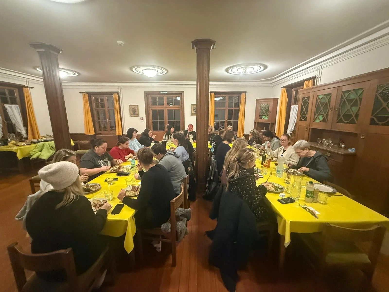 Mehrere Menschen sitzen an einem langen Esstisch mit gelben Tischdecken in einem gemütlichen, holzgetäfelten Raum beim Essen.