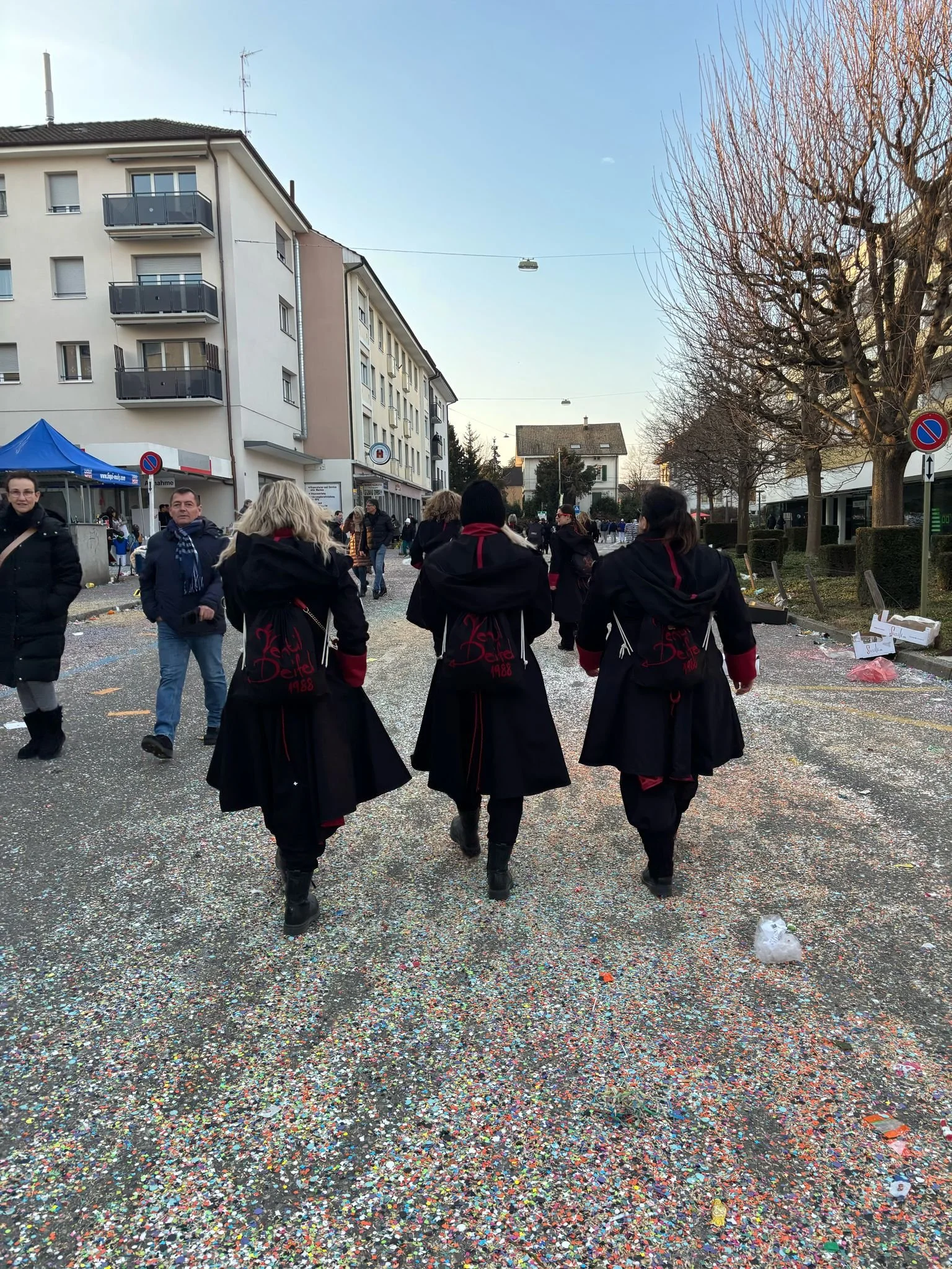Drei Frauen in schwarzen Kostümen mit roten Akzenten, die bei einer Feier oder Parade unterwegs sind, gehen auf einer Straße mit Konfetti und Müll, umgeben von anderen Menschen und Gebäuden, während die Bäume Blätter verloren haben.