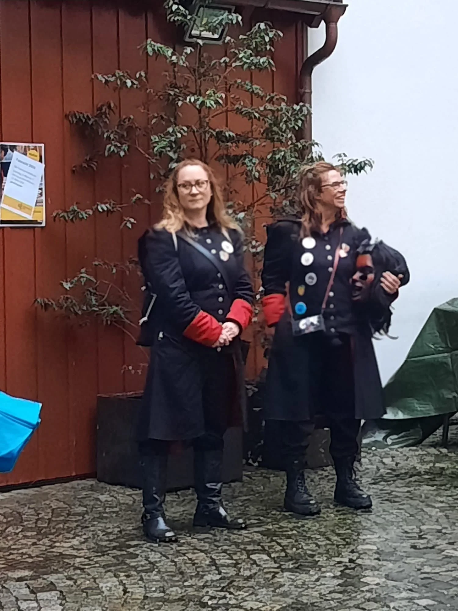 Zwei Frauen stehen nebeneinander vor einer roten Holzwand, tragen schwarze Mäntel mit roten Ärmeln und spenden. Beide sind mit Buttons geschmückt, eine hat eine schwarze Tasche und die andere hält eine schwarze Jacke. Der Hintergrund ist bewölkt, und