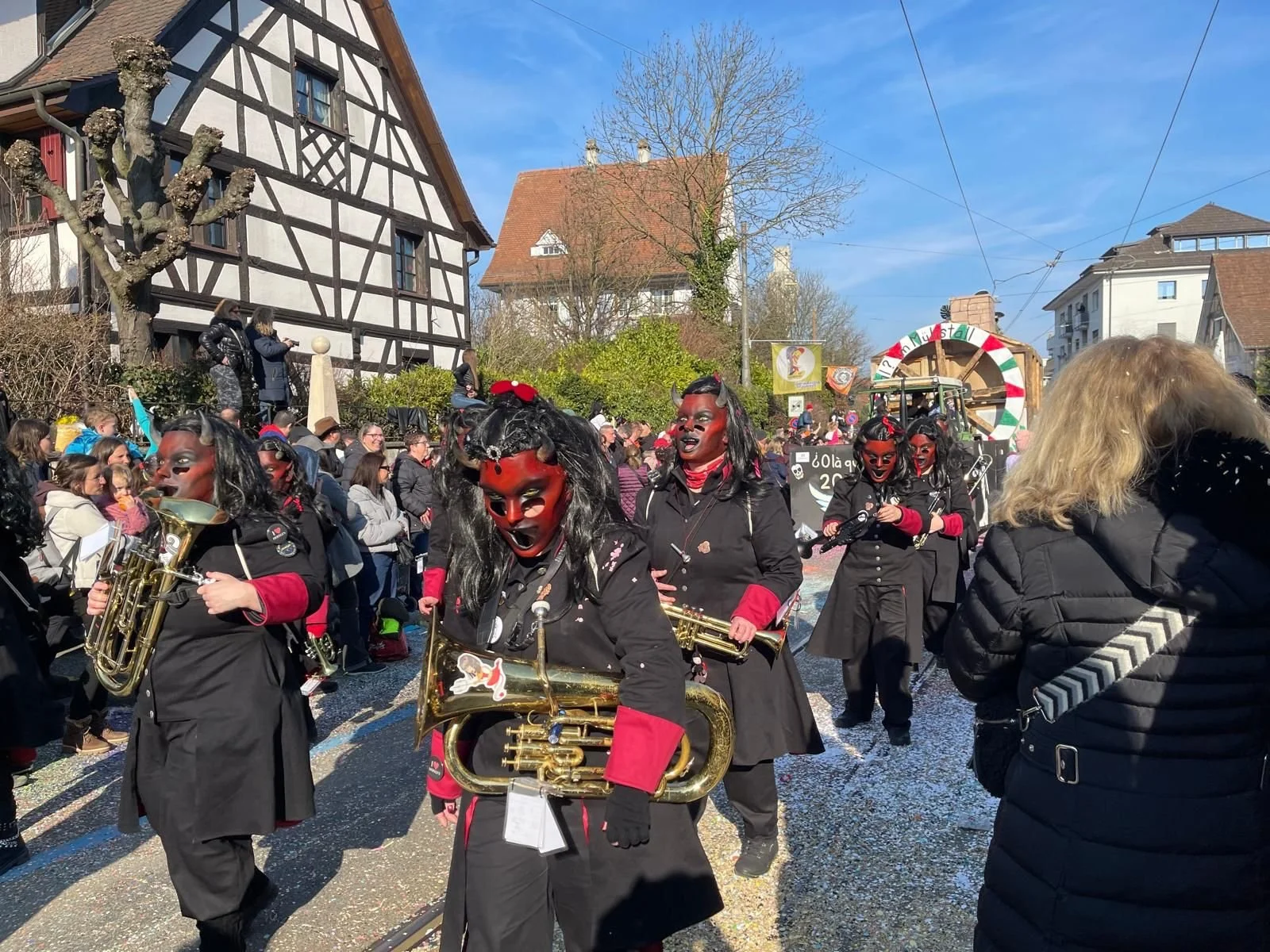Menschen in Kostümen bei einem Karnevalsumzug, einige tragen Teufelskostüme mit roten Masken und Hörnern, andere spielen Musikinstrumente, im Hintergrund ist ein Wagen mit buntem Dekor.