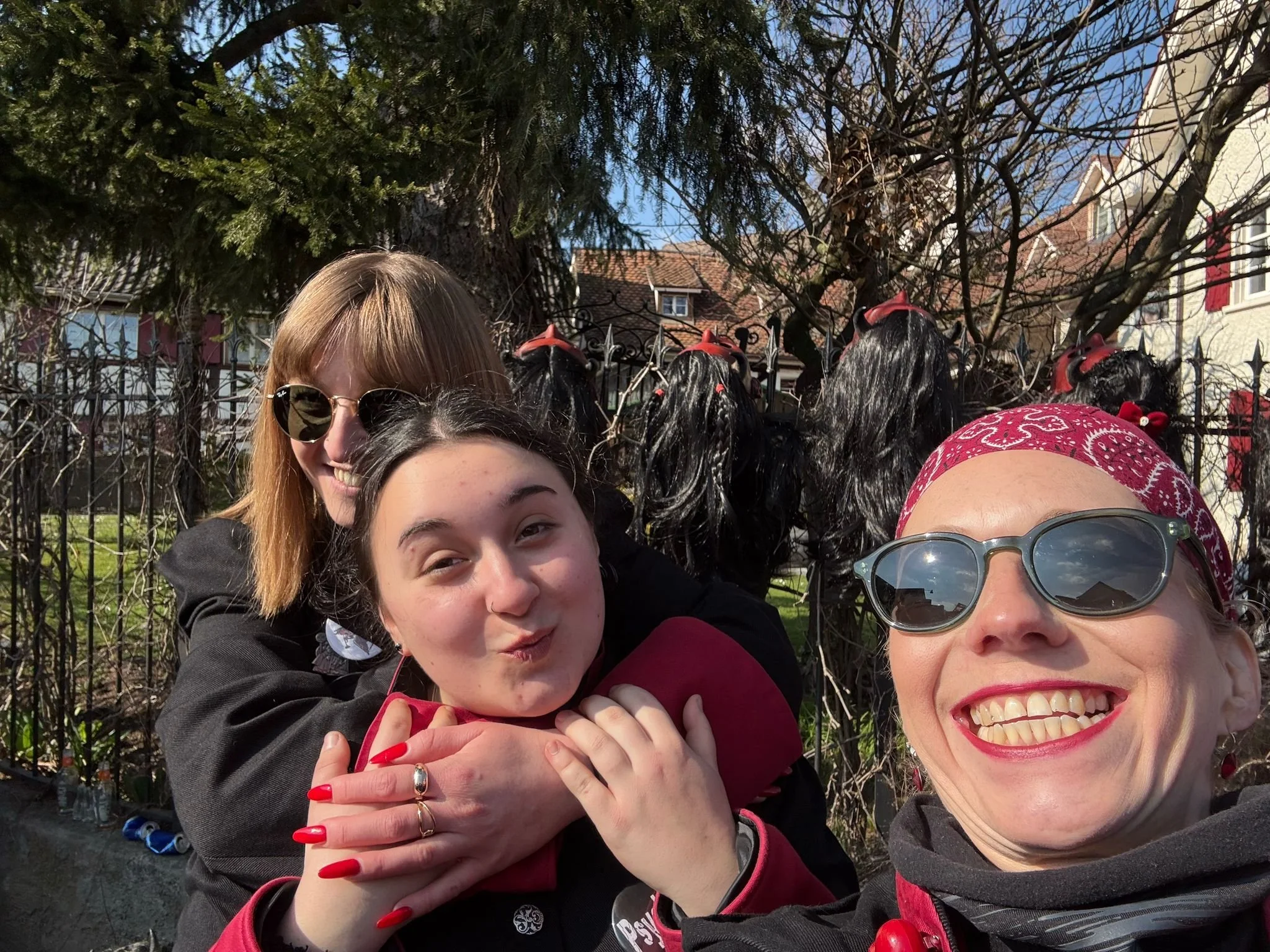 Drei Frauen machen ein Selfie vor einem Zaun mit schwarzen Perücken im Hintergrund, bei sonnigem Wetter.