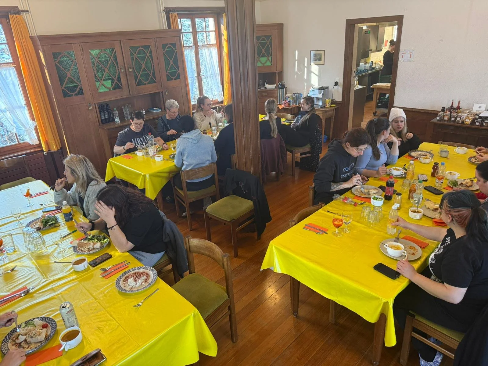 Mehrere Menschen sitzen an Tischen mit gelben Tischdecken und essen in einem rustikalen Restaurant mit Holzboden und Holzinterieur.