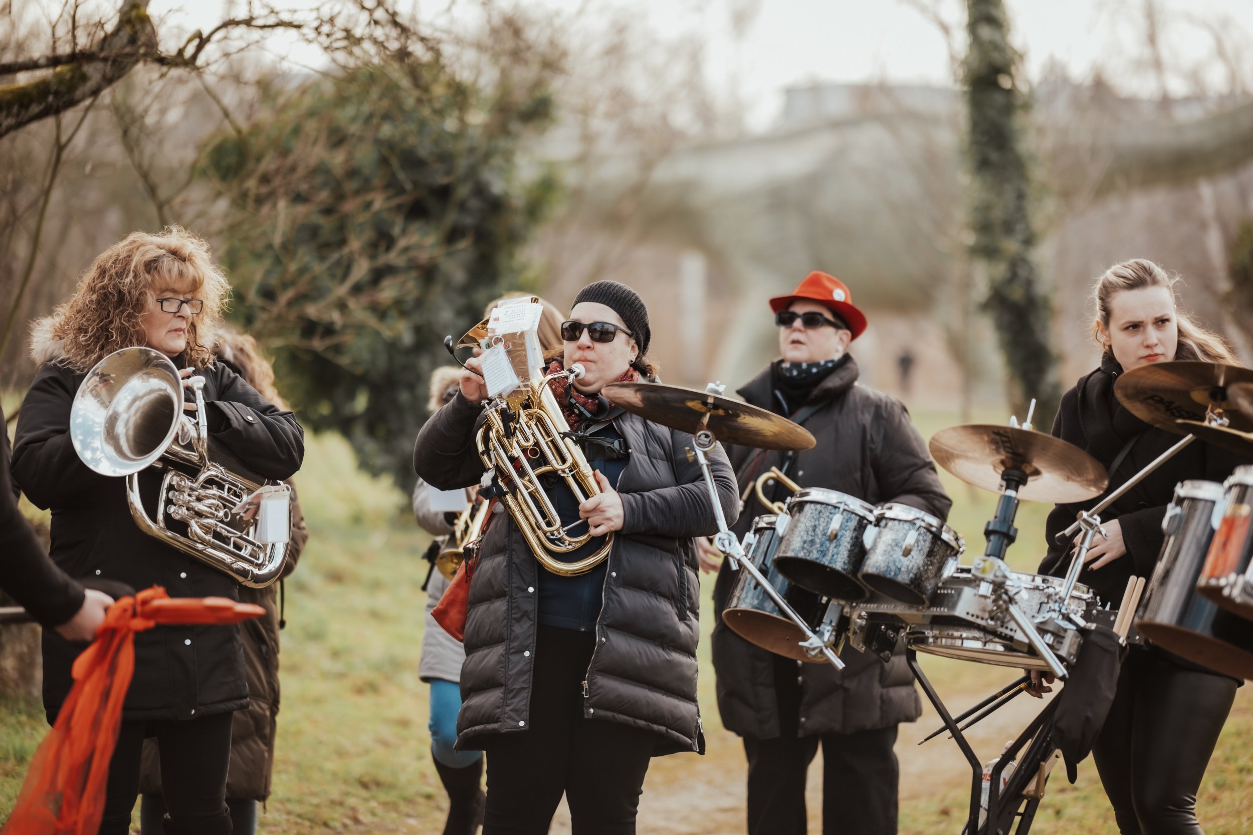 Gruppe von Musikern draußen in einem Park, die verschiedene Instrumente spielen, inklusive Trompete, Snare-Drum, und elektronisches Schlagzeug, bei kühlem Wetter mit Bäumen im Hintergrund.