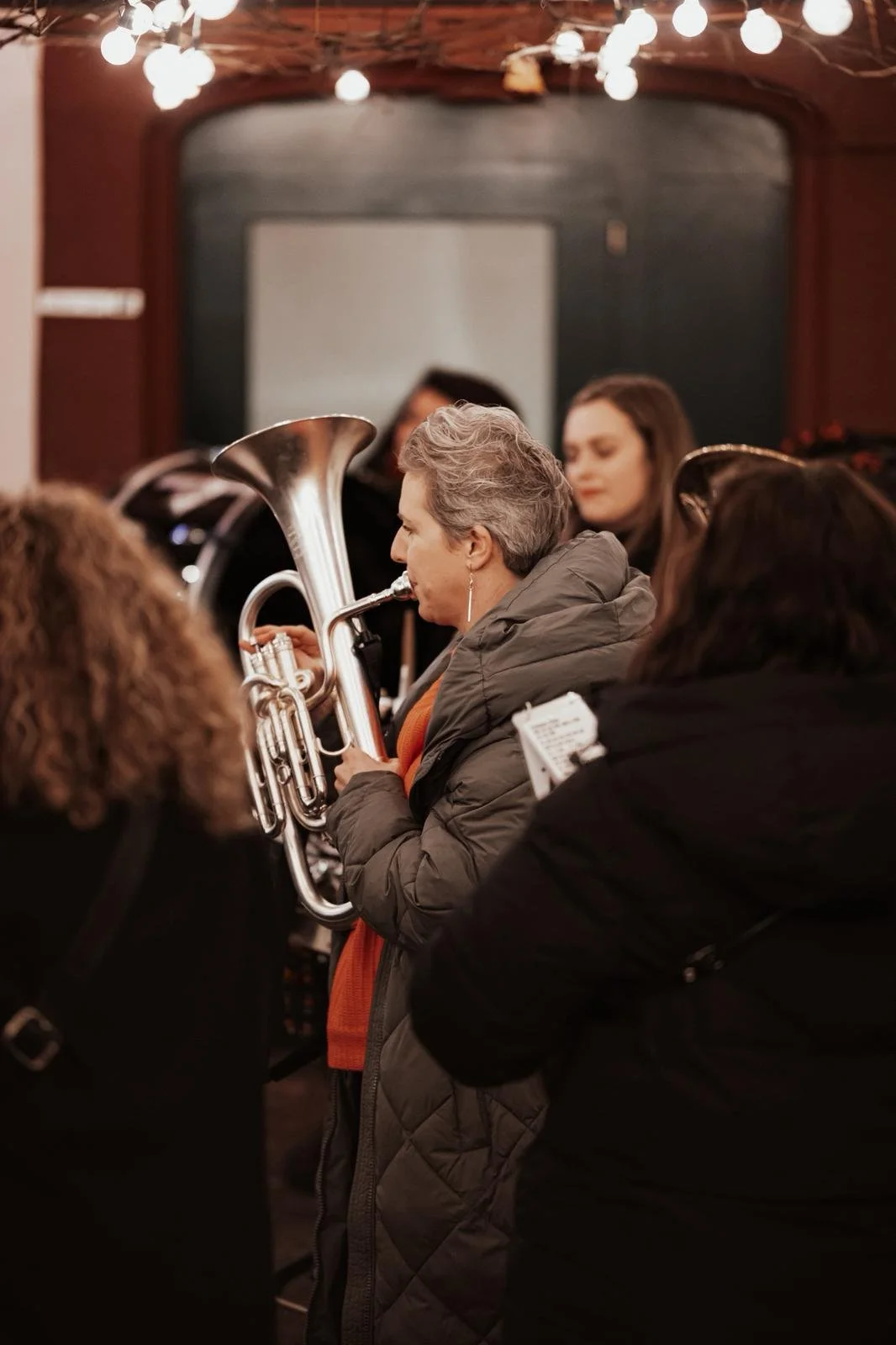 Eine Frau mit grauen Haaren, die ein Blasinstrument spielt, umgeben von anderen Menschen in einem Raum mit Laternen an der Decke.