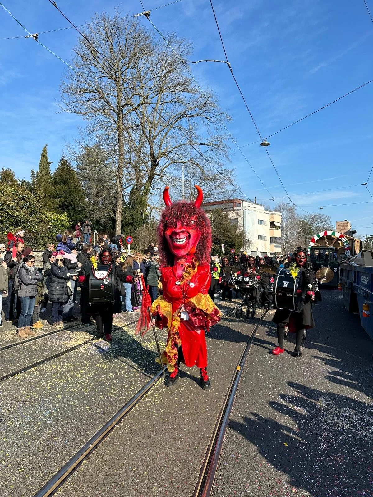 Karnevalsumzug mit mehreren geschminkten und kostümierten Menschen, ein großer Teufelsmaskenläufer in rotem Kostüm und Teufelshörnern in der Mitte, bunte Konfetti auf der Straße, im Hintergrund Bäume und Häuser, blauer Himmel.