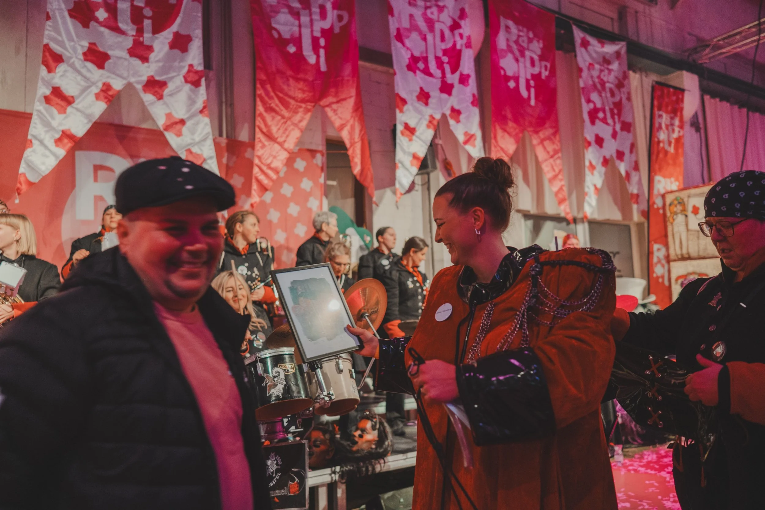 Menschen feiern bei einer Veranstaltung mit roten und weißen Bannern im Hintergrund, einige lachen und unterhalten sich, im Vordergrund eine lachende Frau, die ein Tablet hält.