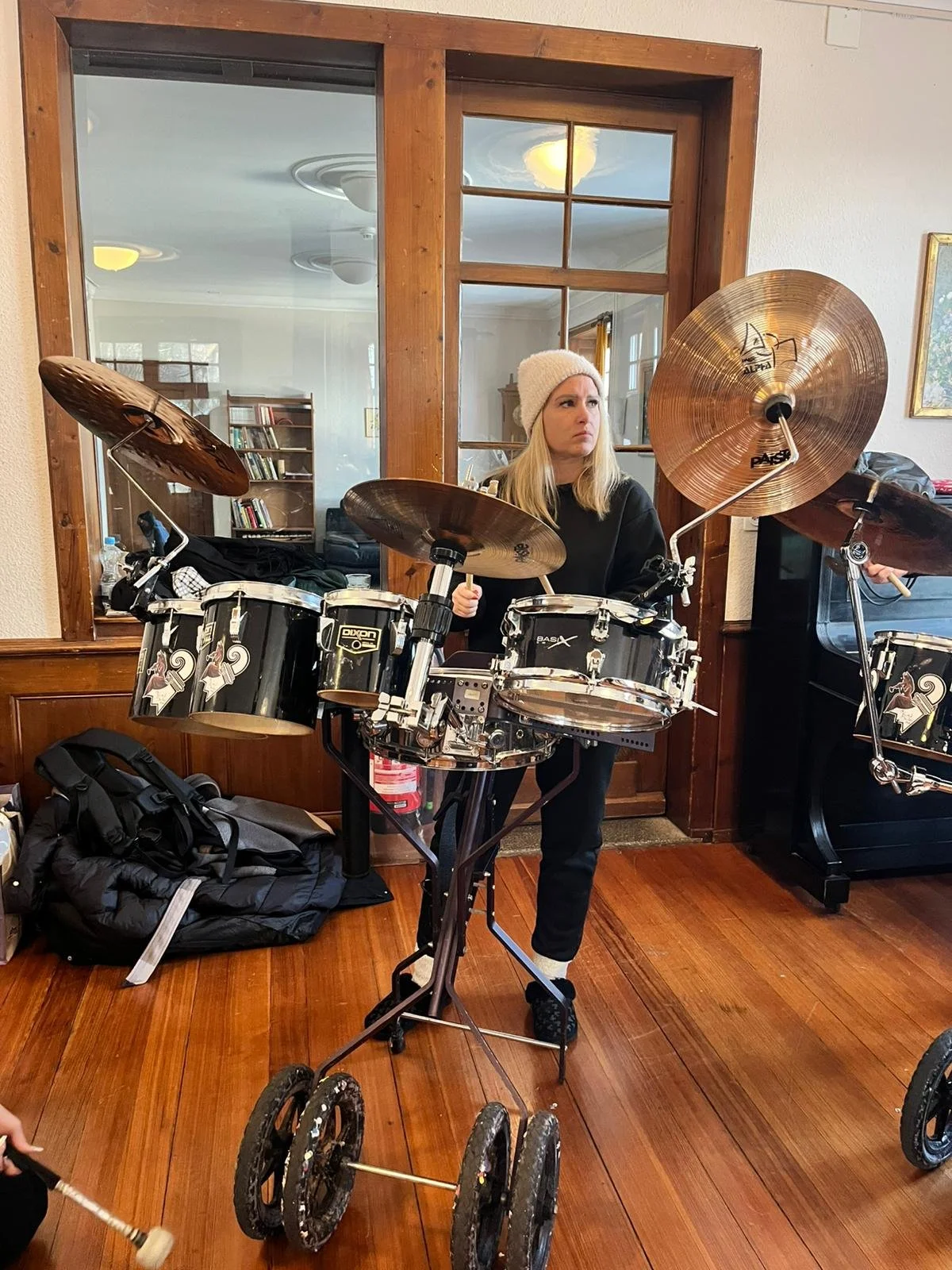 Junge Frau mit langem blondem Haar, weißer Mütze und schwarzer Kleidung sitzt hinter einem als Drumset umgebauten Rollstuhl mit Musikinstrumenten, im Raum mit Holzboden, Fensterwand und Wegen im Hintergrund.