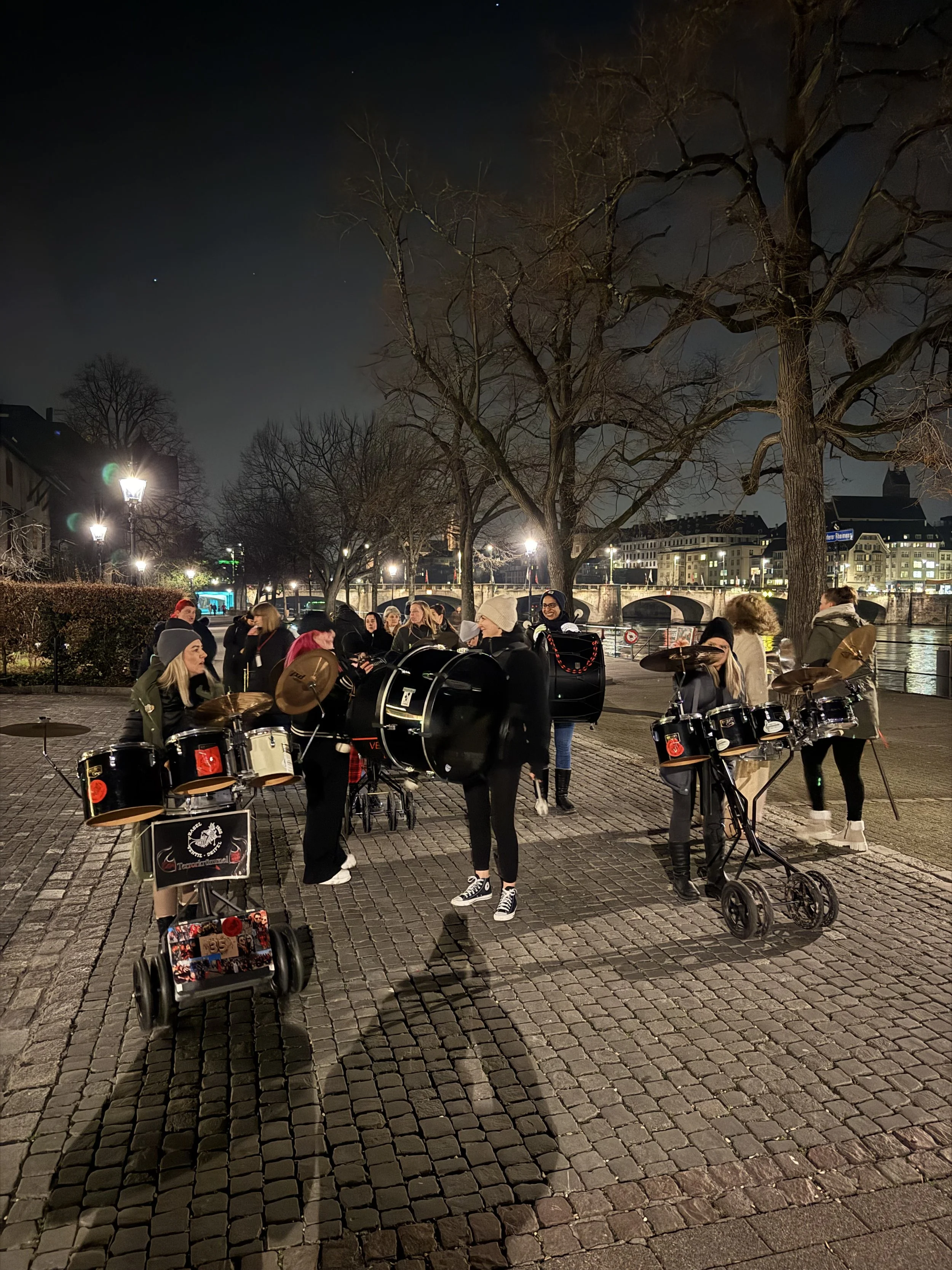 Gruppe von Menschen mit Musikinstrumenten, die nachts an einem Flussufer in einer Stadt stehen, umgeben von Bäumen ohne Blätter, beleuchtet durch Straßenlampen.