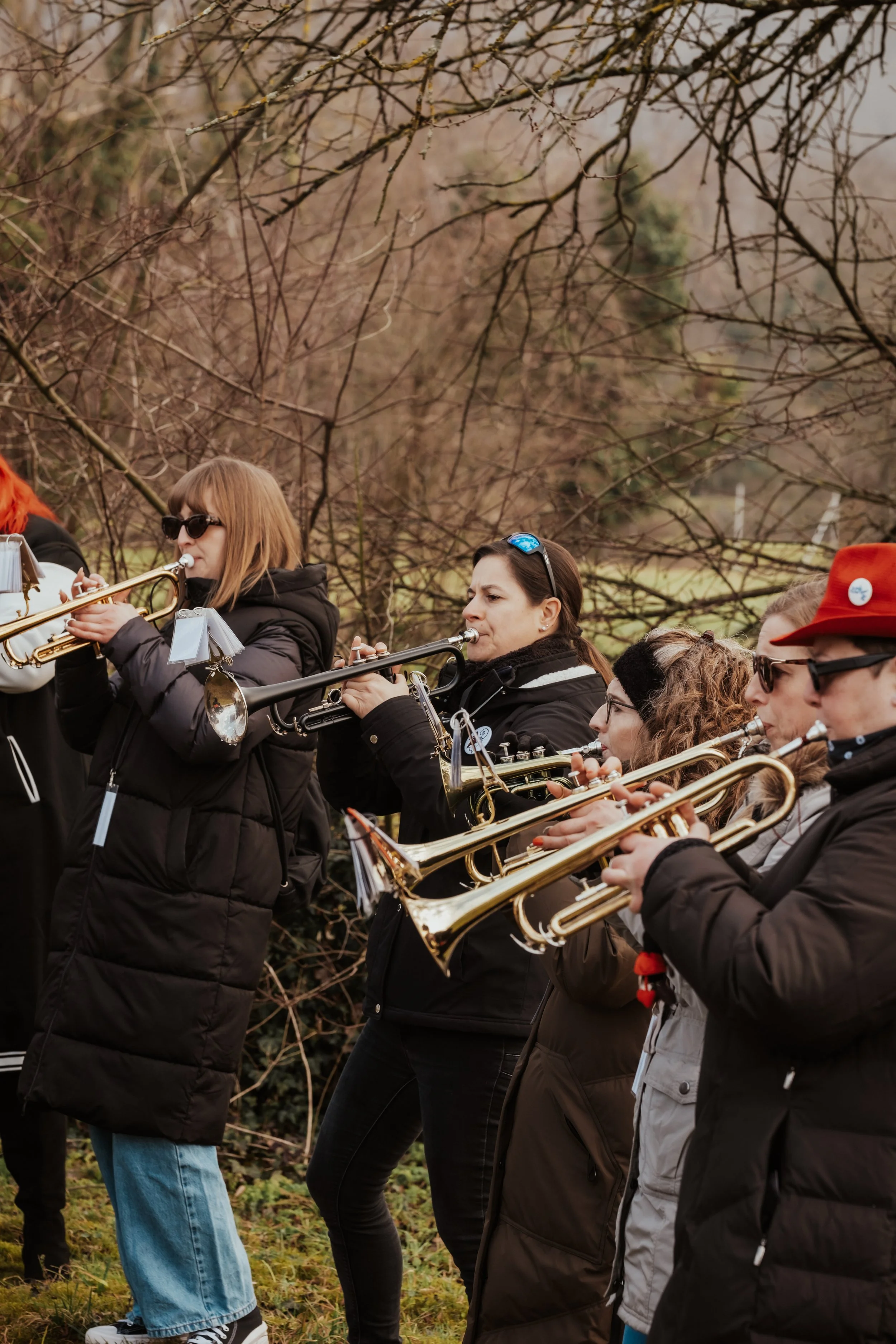 Gruppe von Musikerinnen spielt Blasinstrumente im Freien, umgeben von Bäumen ohne Blätter bei wechselhaftem Wetter.