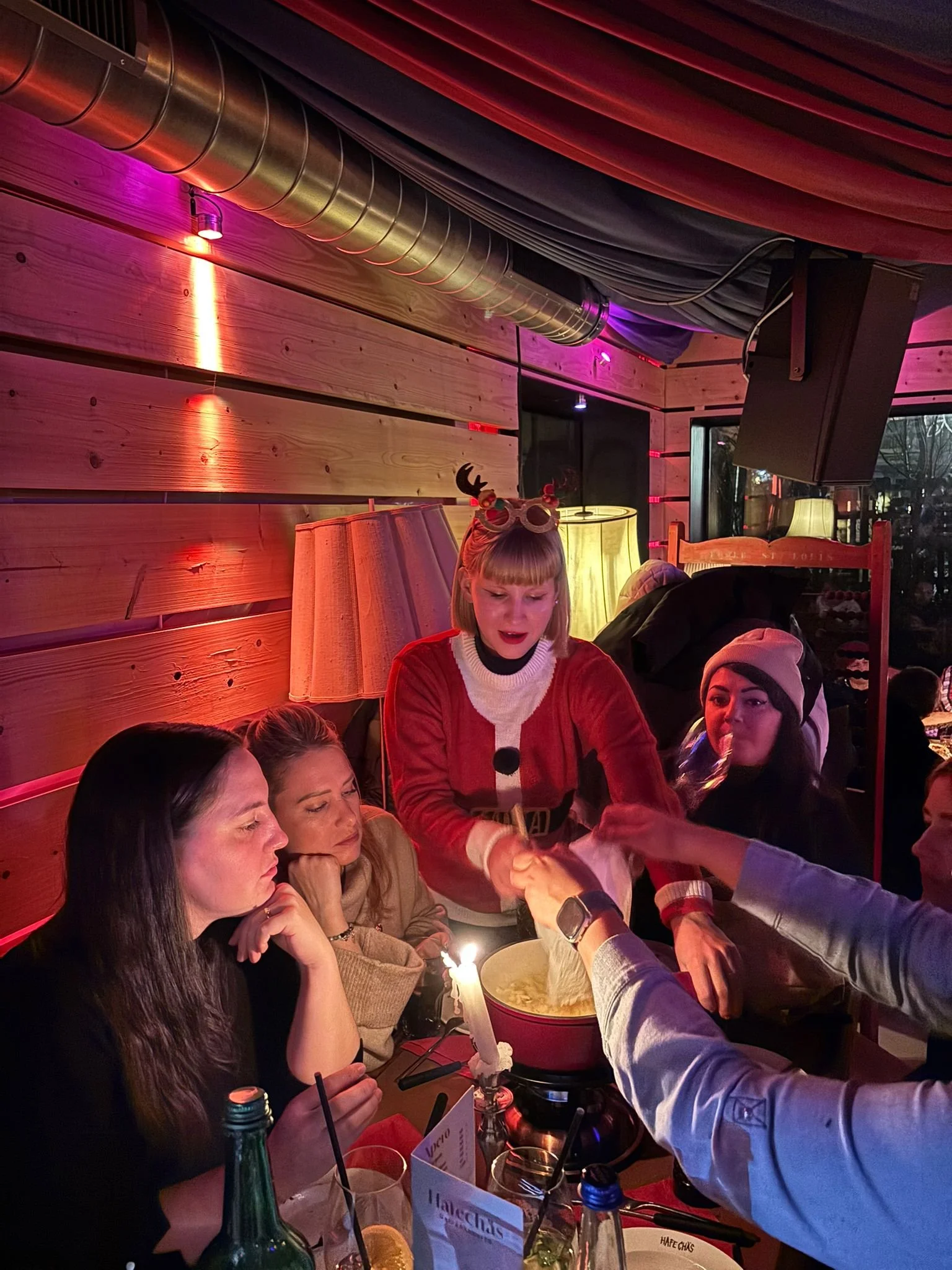 Eine Gruppe von Freunden bei einem Abendessen in einem Restaurant, die eine festliche Stimmung haben. Es sind vier Frauen, und eine Frau mit einem Weihnachtskostüm und Rentier-Ohren stopft Essen in eine Schüssel, während die anderen zuschauen. Die Ti