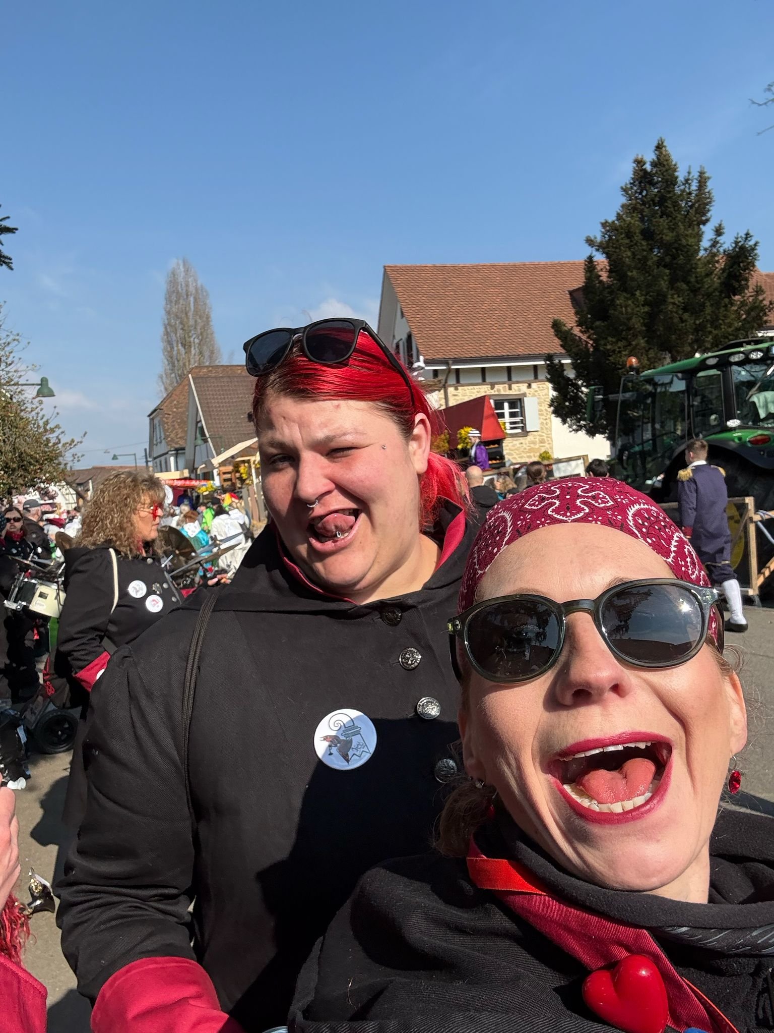 Zwei lachende Frauen mit dunklen Sonnenbrillen und roten Accessoires posieren auf einem Fest mit vielen Menschen, Gebäuden und einem Traktor im Hintergrund bei schönem Wetter.