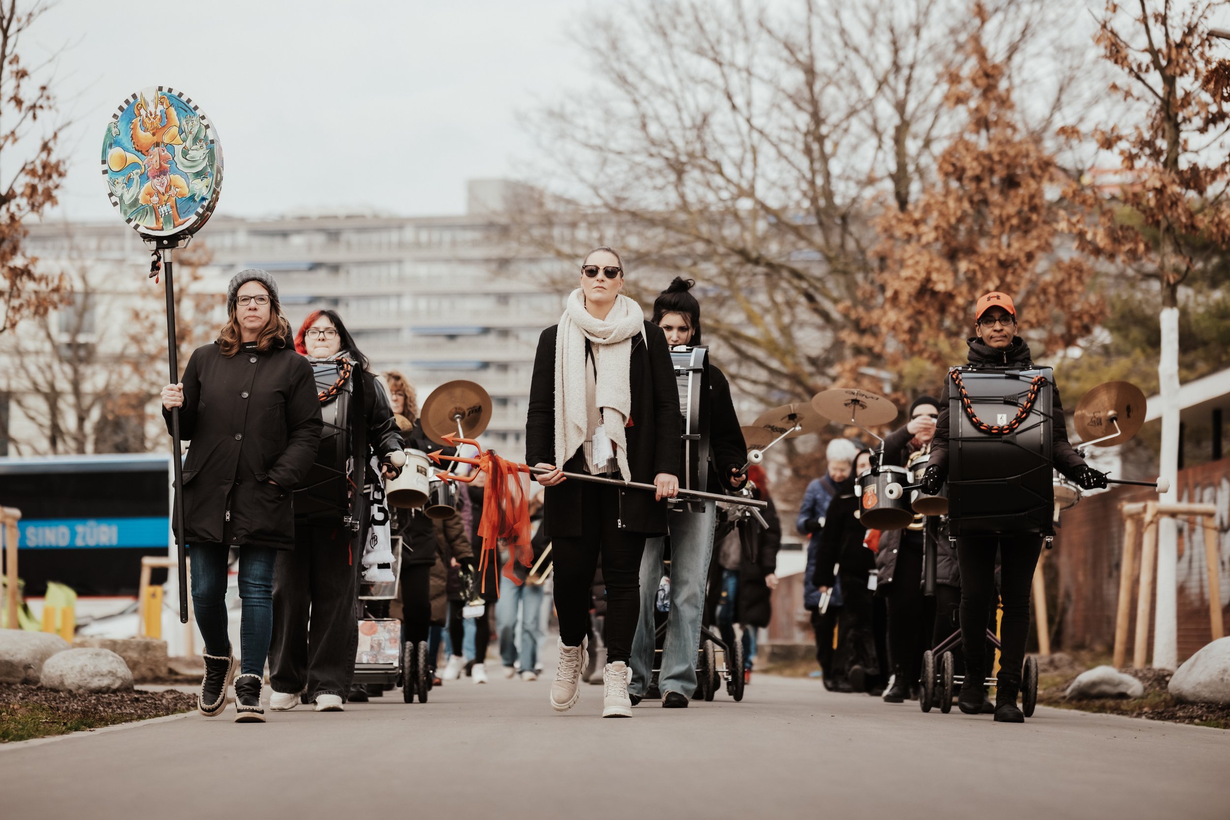 Gruppe von Menschen, die in einer Demonstration marschieren, einige tragen Musikinstrumente und ein Ballon, mit herbstlichen Bäumen im Hintergrund.