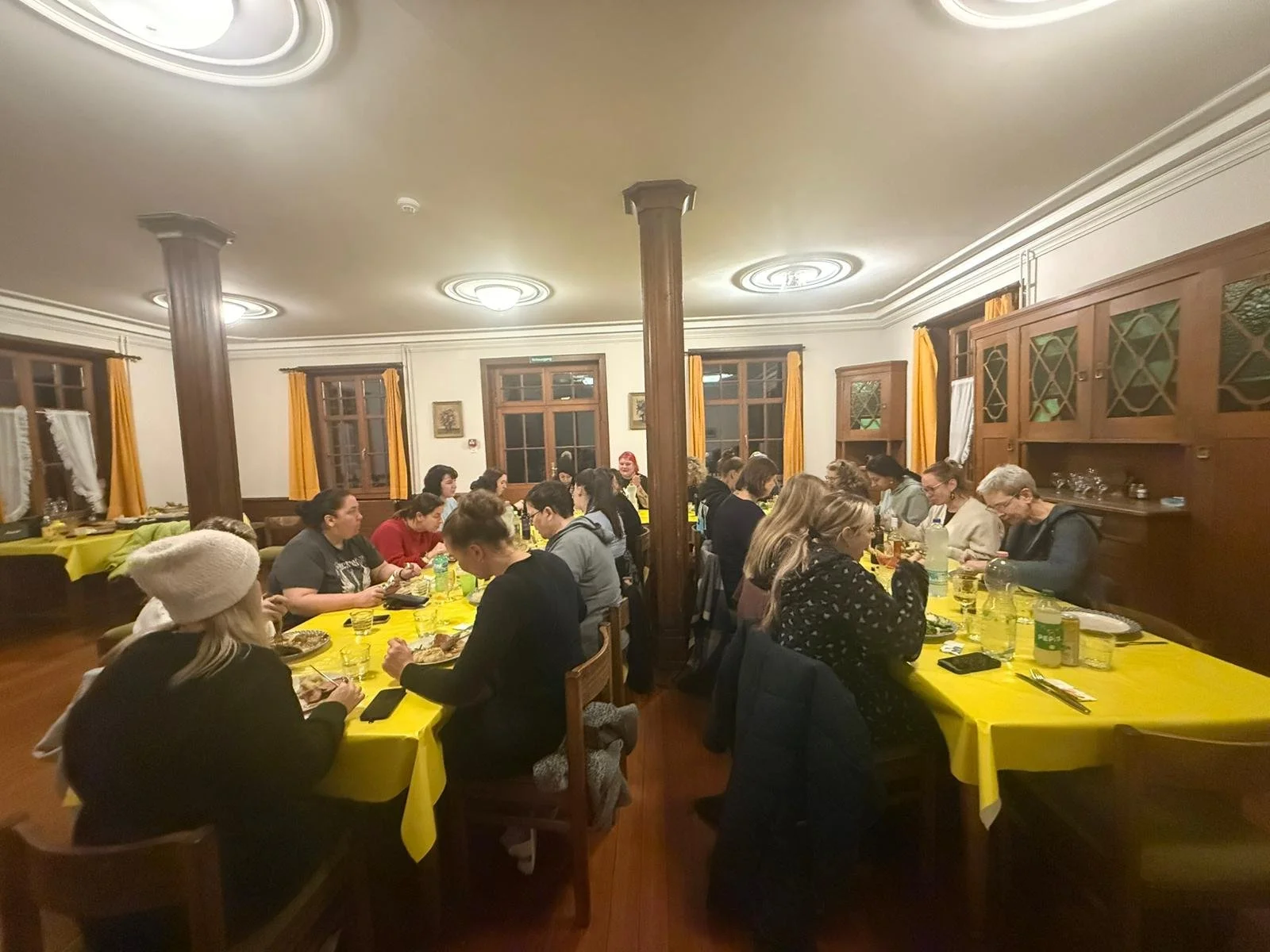 Mehrere Menschen sitzen an zwei Tischen mit gelben Tischtüchern, essen und trinken in einem gut beleuchteten Esszimmer mit Holzvertäfelung und gelben Vorhängen.