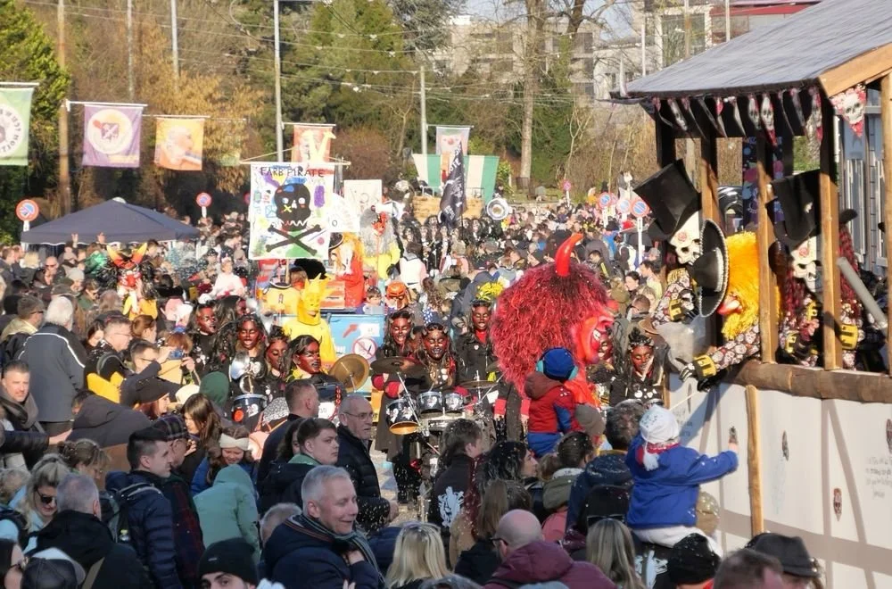 Bunte Karnevalsparade mit Menschen in Kostümen, Musikanten, großen Figuren und dekorativen Bannern bei Sonnenschein.