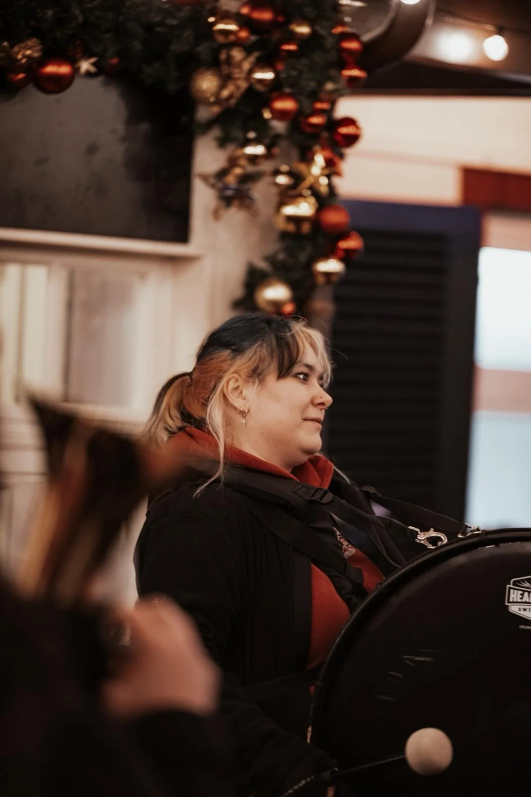Eine Frau mit blondem und braunem Haar, die an einem Tisch in einem Raum mit Weihnachtsdekoration sitzt, im Hintergrund ein Weihnachtsbaum mit Kugeln. Die Frau schaut nach rechts, trägt Ohrringe und einen dunklen Pullover.