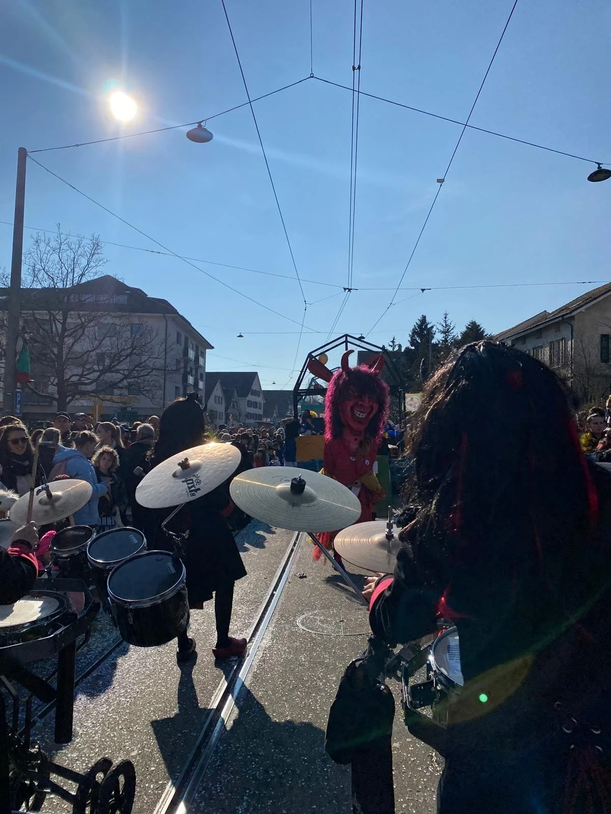 Menschen feiern auf einer Straße mit Musik und Kostümen bei sonnigem Wetter. Ein Musiker spielt Trommeln, im Hintergrund eine Person in einem Teufelskostüm.
