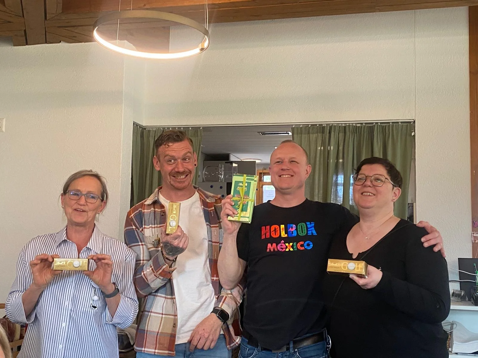 Vier Personen, zwei Männer und zwei Frauen, halten Goldbarren und lachen in einem Raum mit grünen Vorhängen im Hintergrund.