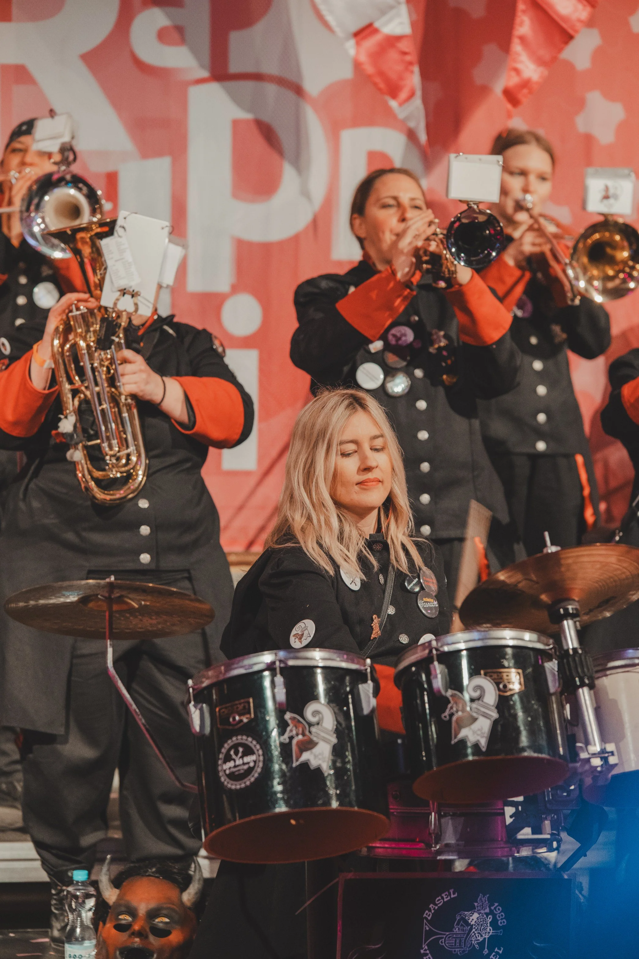 Musikerinnen spielen ein Blech- und ein Schlagzeug bei einer Bühneleistung, alle tragen schwarze Uniformen mit roten Akzenten