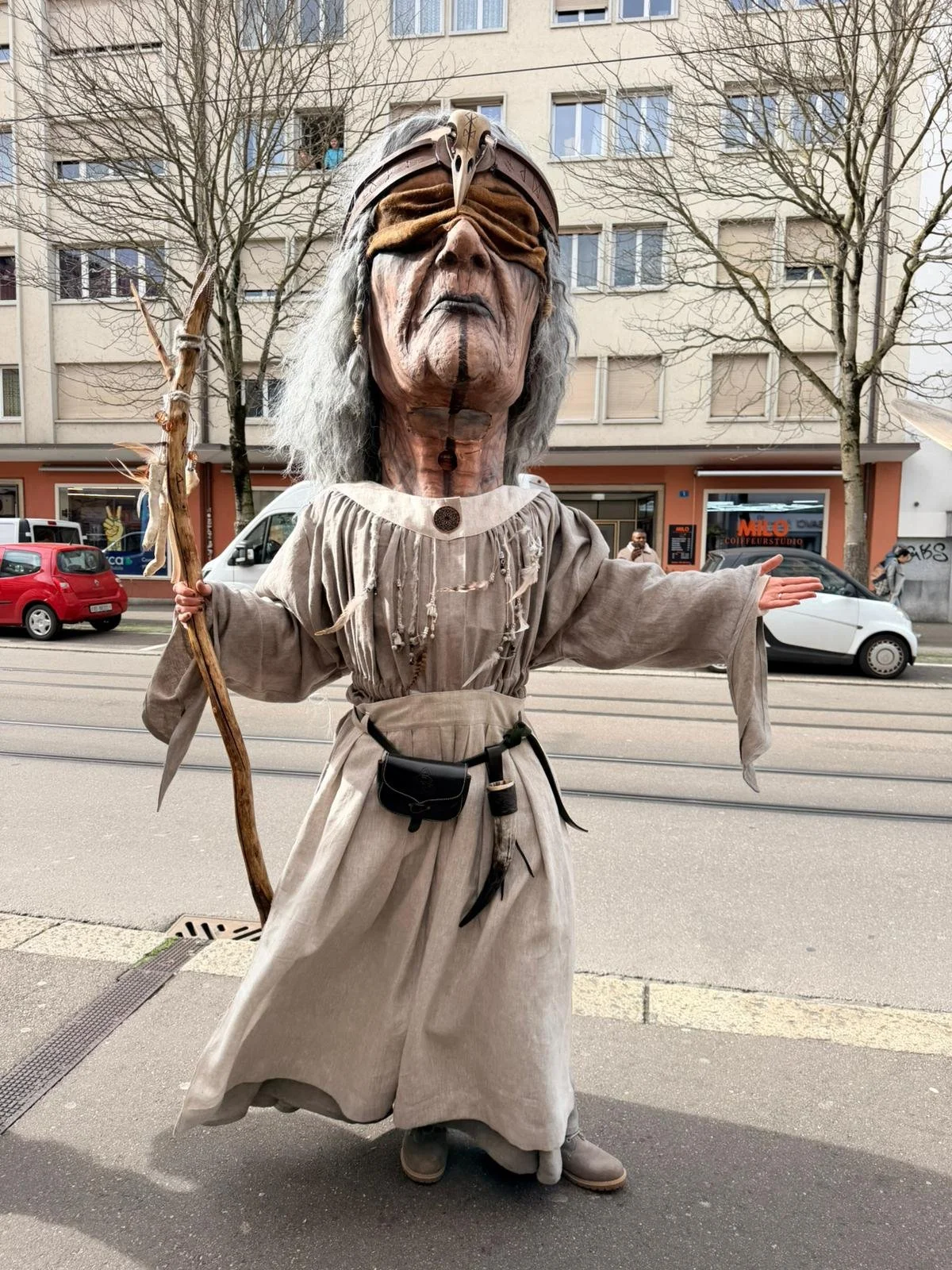 Eine Person trägt eine große, detaillierte Kostümmaske eines alten, weisen Zauberers mit grauem Haar und braunem Hemd. Der Zauberer hält einen Stock in der Hand und steht auf einer Straßenseite vor einem Stadtgebäude.