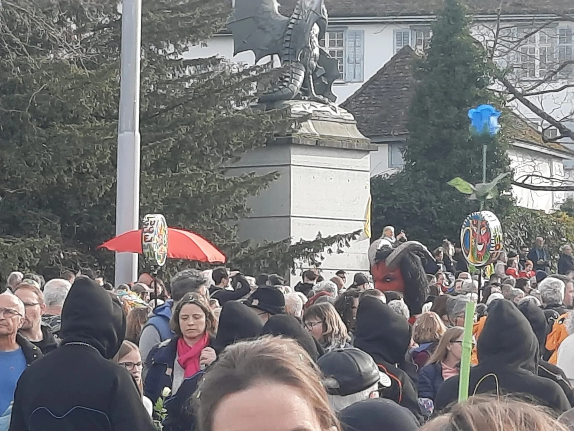 Menschen versammeln sich vor einer Statue in einem öffentlichen Platz, einige halten bunte Schilder und Schirme, im Hintergrund sind Bäume und mehrstöckige Gebäude sichtbar.