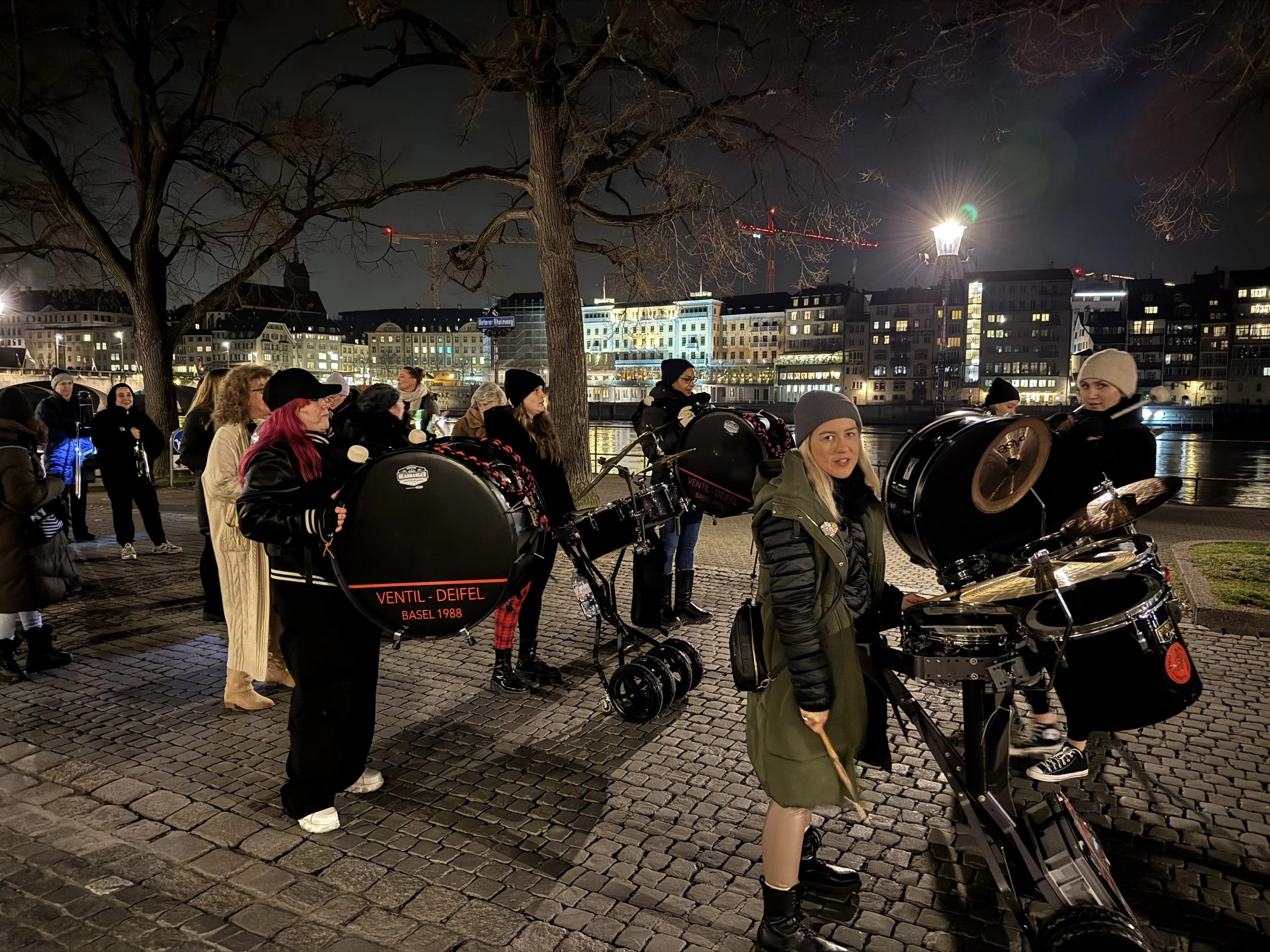Gruppe von Musikern mit Schlagzeugen und Tretrollern am Flussufer bei Nacht, beleuchtet durch Straßenlaternen, in einer Stadt mit Gebäuden und Kräne im Hintergrund.