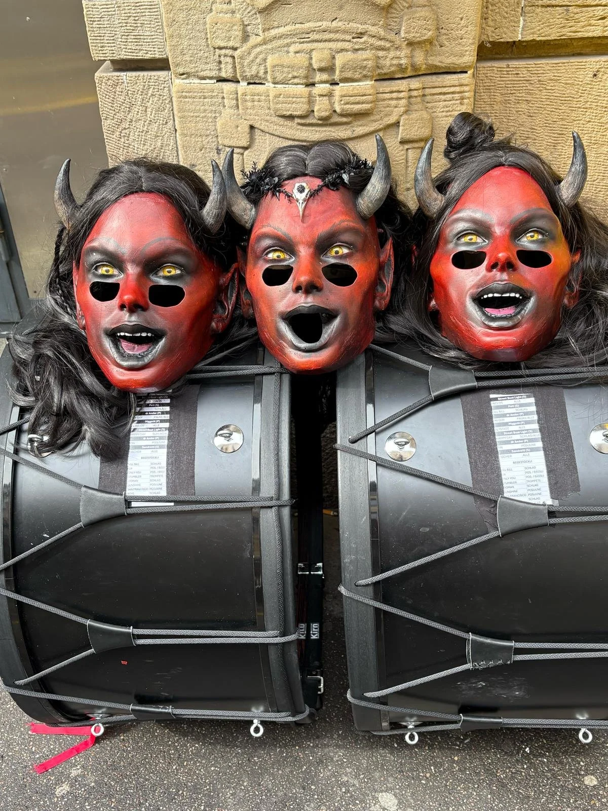 Drei Dämonen-Masken mit roten Gesichtern, schwarzen Hornplatten, gelben Augen und offenen Mündern auf schwarzen Karren.