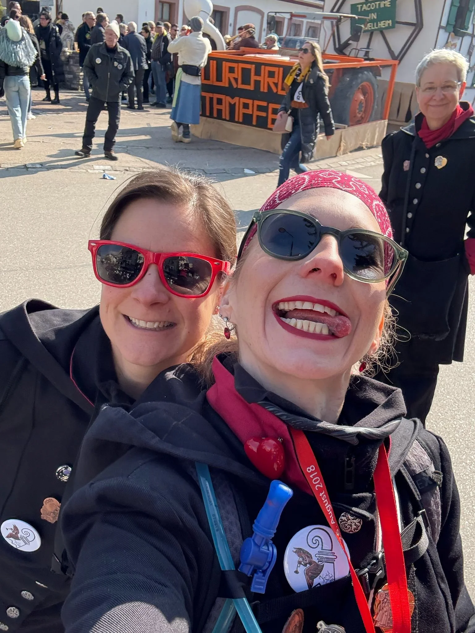 Zwei lachende Frauen tragen Sonnenbrillen und sind auf einem Straßenfest, während im Hintergrund Menschen und eine Parade mit einem Wagen zu sehen sind