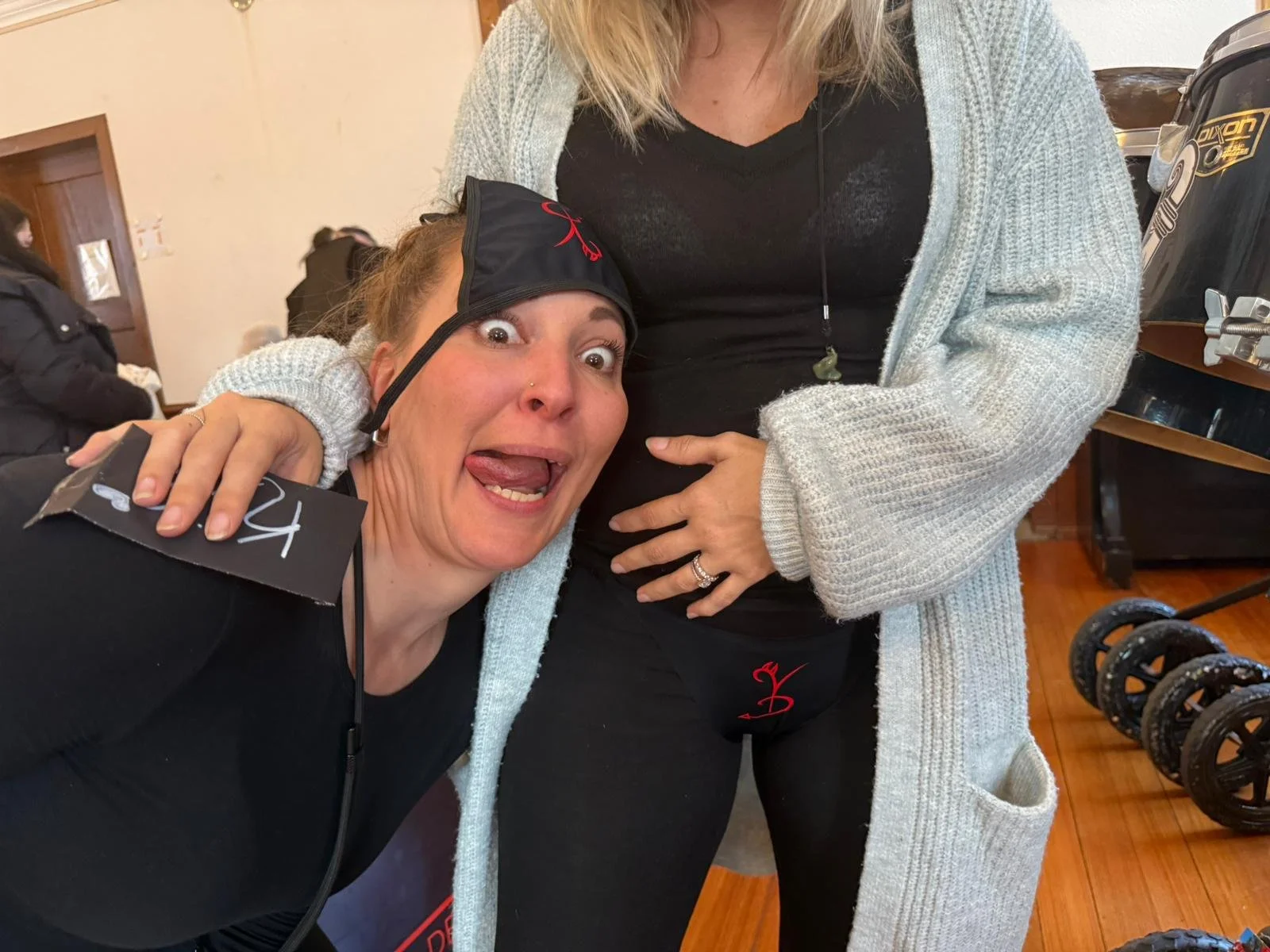 Zwei Frauen, eine ist lachend und macht ein witziges Gesicht, trägt eine schwarze Mütze, die auf der Schulter einer anderen Frau liegt. Die andere Frau trägt einen grauen Pullover. Sie sind wahrscheinlich in einem Fitnessstudio oder einer Sportumgebu