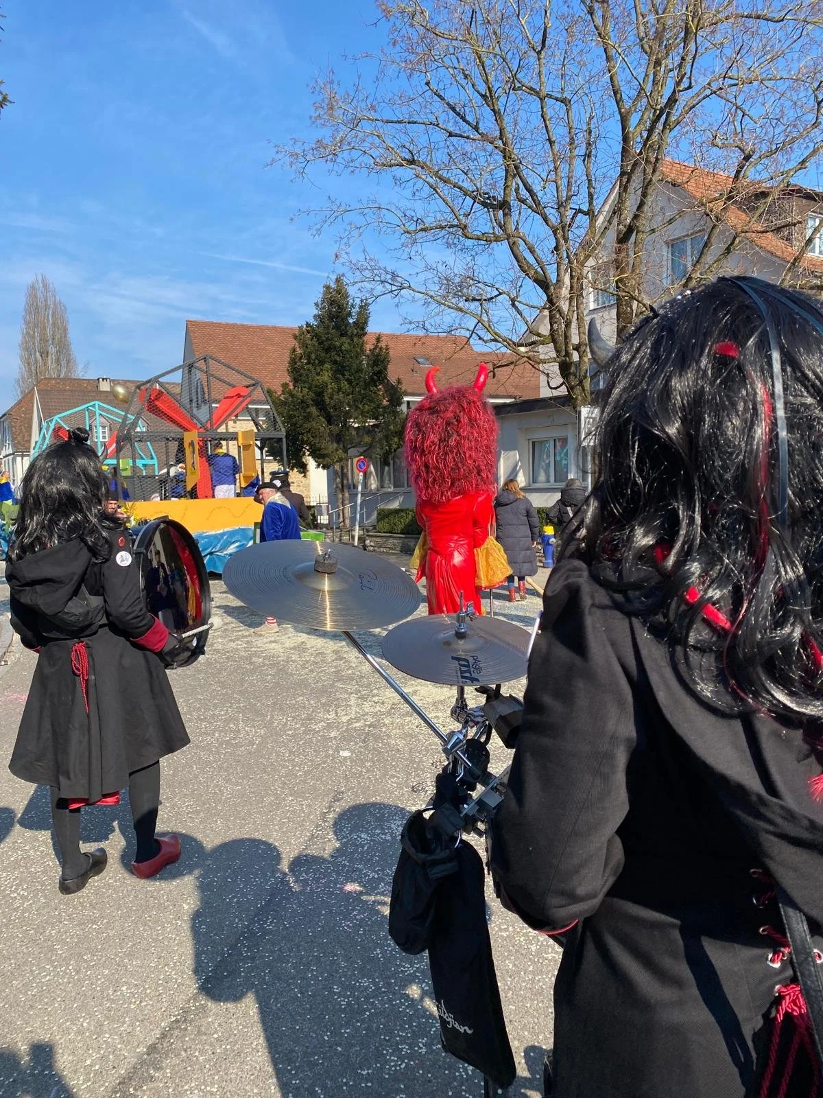 Menschen in Kostümen beim Karneval, vorne eine Person mit schwarzem Haar, rotem Akzent und Trommel, in der Mitte eine Figur mit rotem Anzug, roten Hörnern und rotem Haar, im Hintergrund ein Karnevalswagen mit Rutschbahn und weiteren Menschen im Kostü