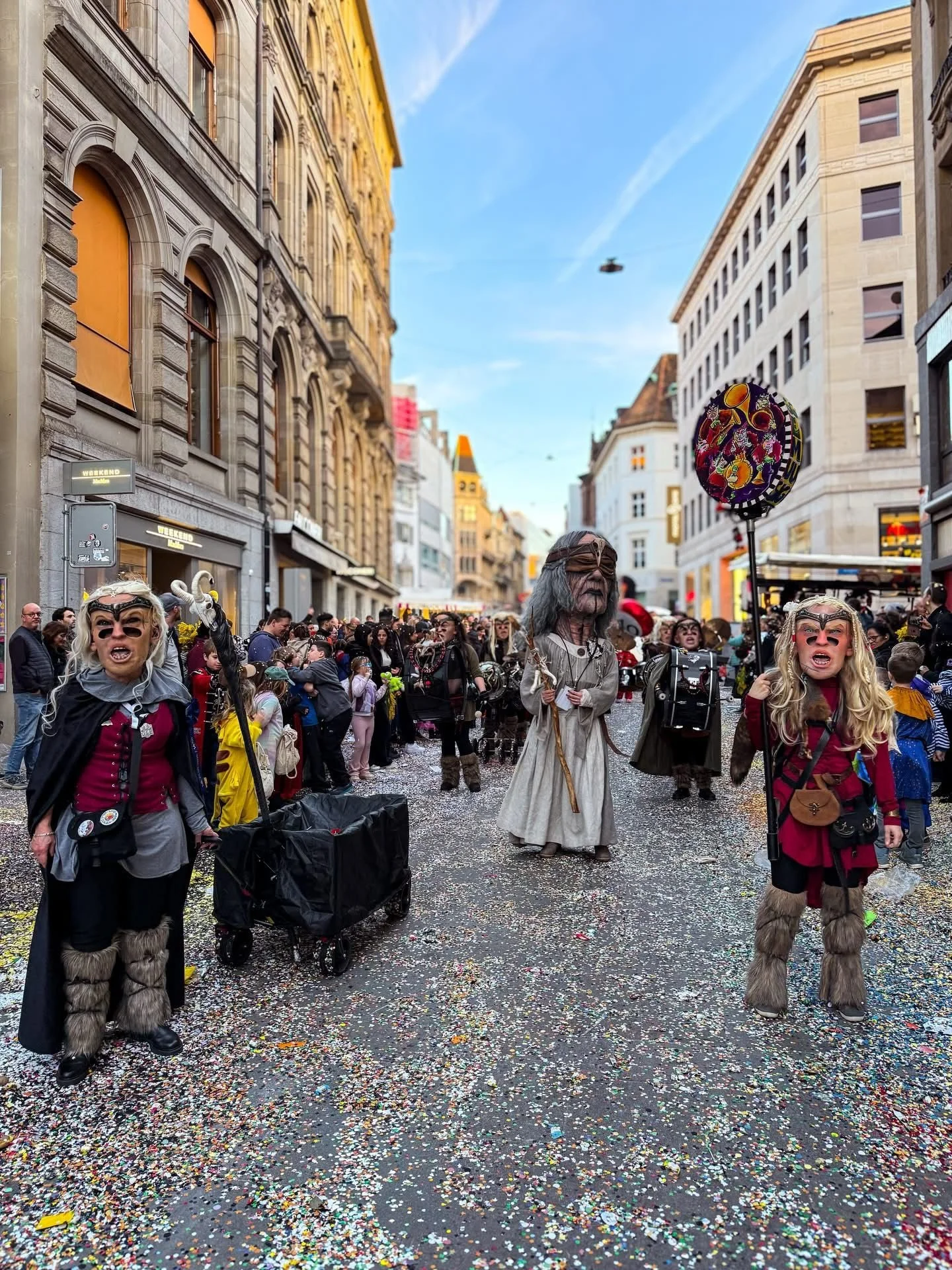 Menschen in Kostümen bei einem Straßenfest, einer trägt eine große Maske, andere halten einen Ballon, überall Konfetti auf dem Boden, im urbanen Stadtzentrum bei blauem Himmel.